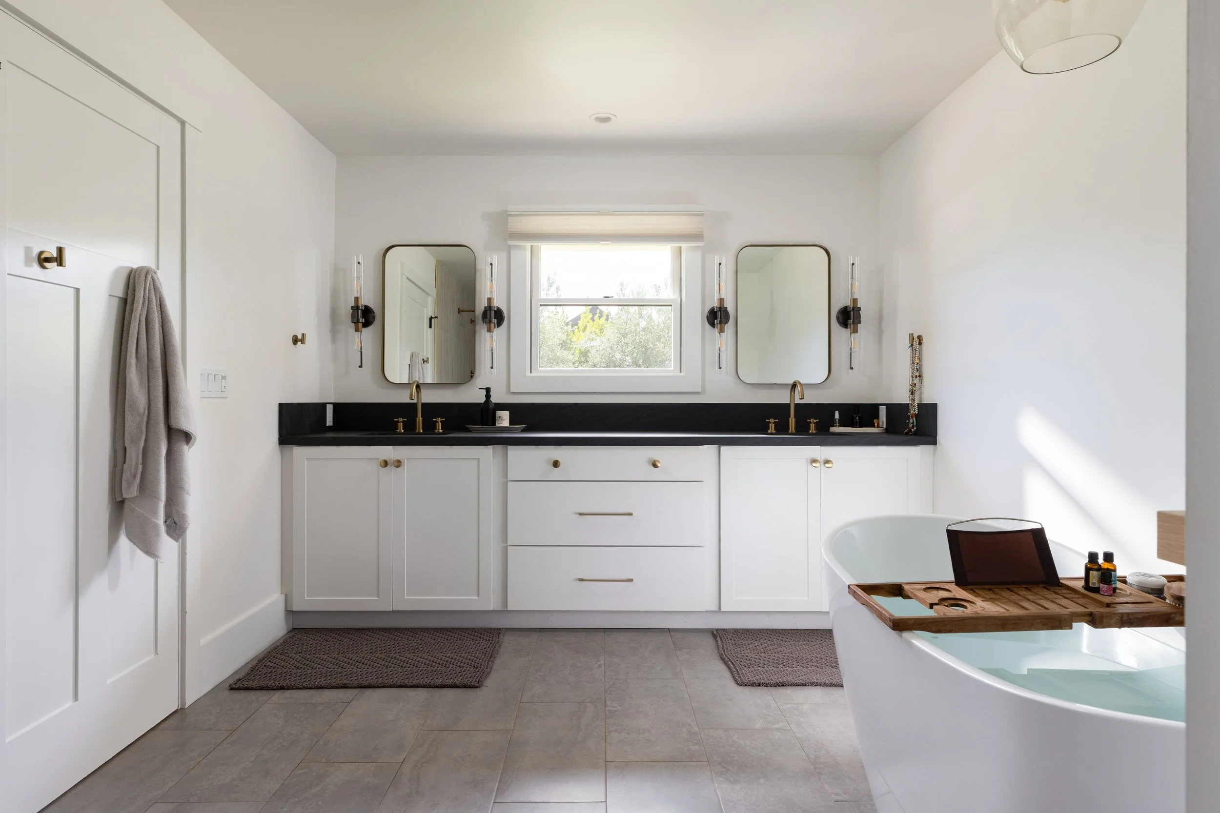 Modern bathroom with white cabinets, black countertop, two mirrors, and gold fixtures. A window above the sink lets in natural light. There are towels on a hook on the left and a wooden tray with toiletries on the right beside a bathtub.