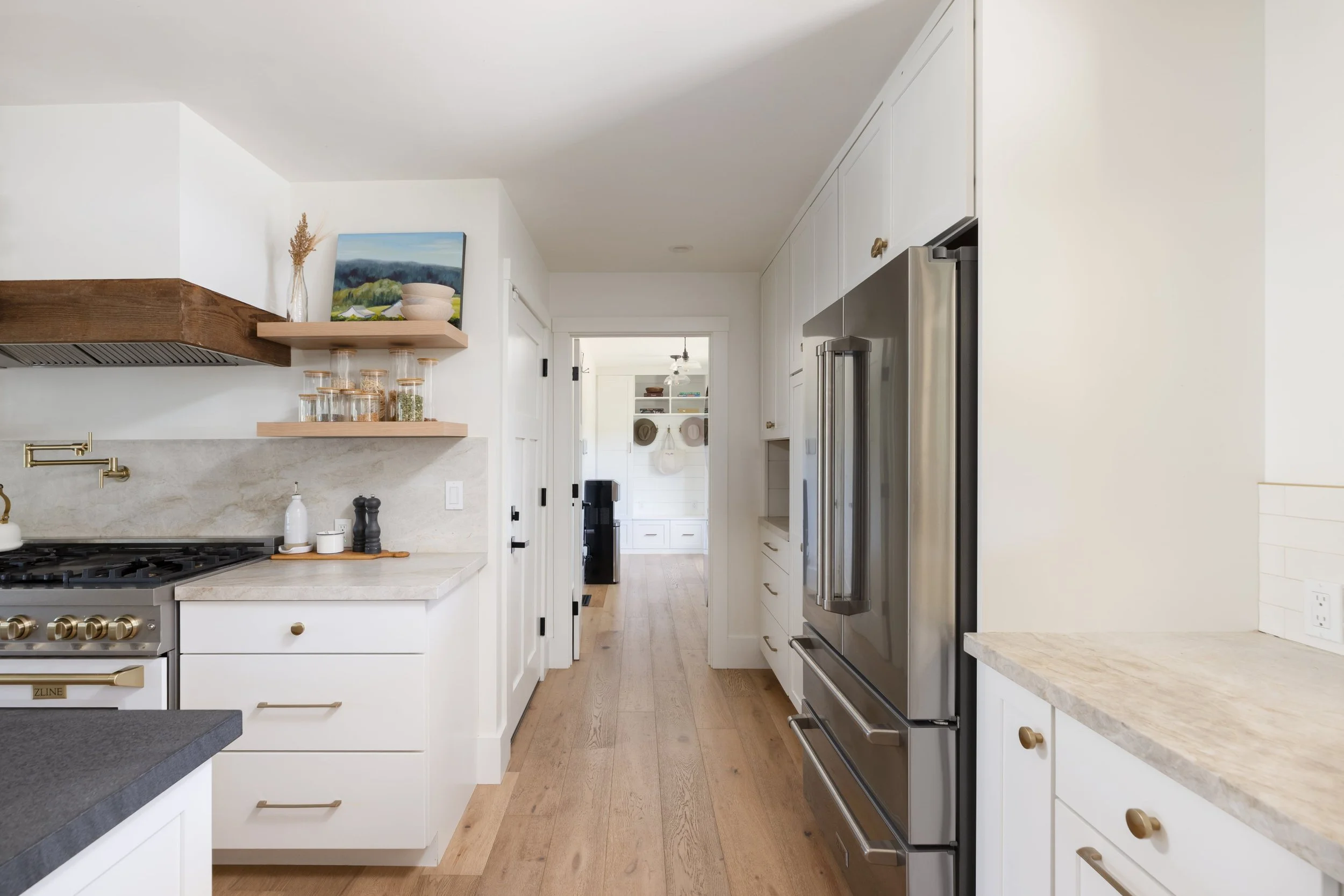 Modern kitchen with white cabinets, stainless steel appliances, and light wood flooring, view down the hallway to a room with shelves.