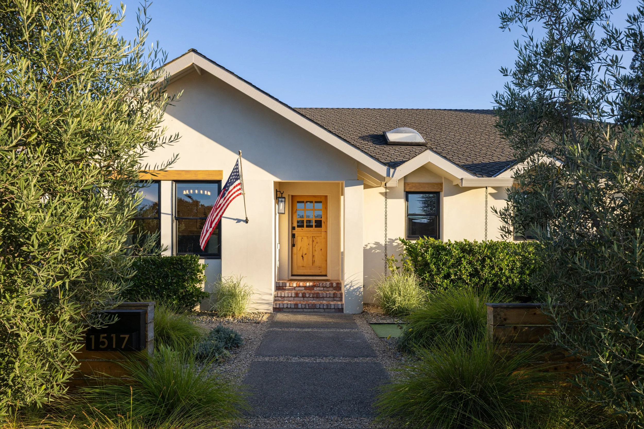 Front view of a house with a gabled roof, beige exterior walls, a wooden front door, and a small set of brick steps, surrounded by green bushes and trees, with an American flag hanging near the door, under a clear blue sky.