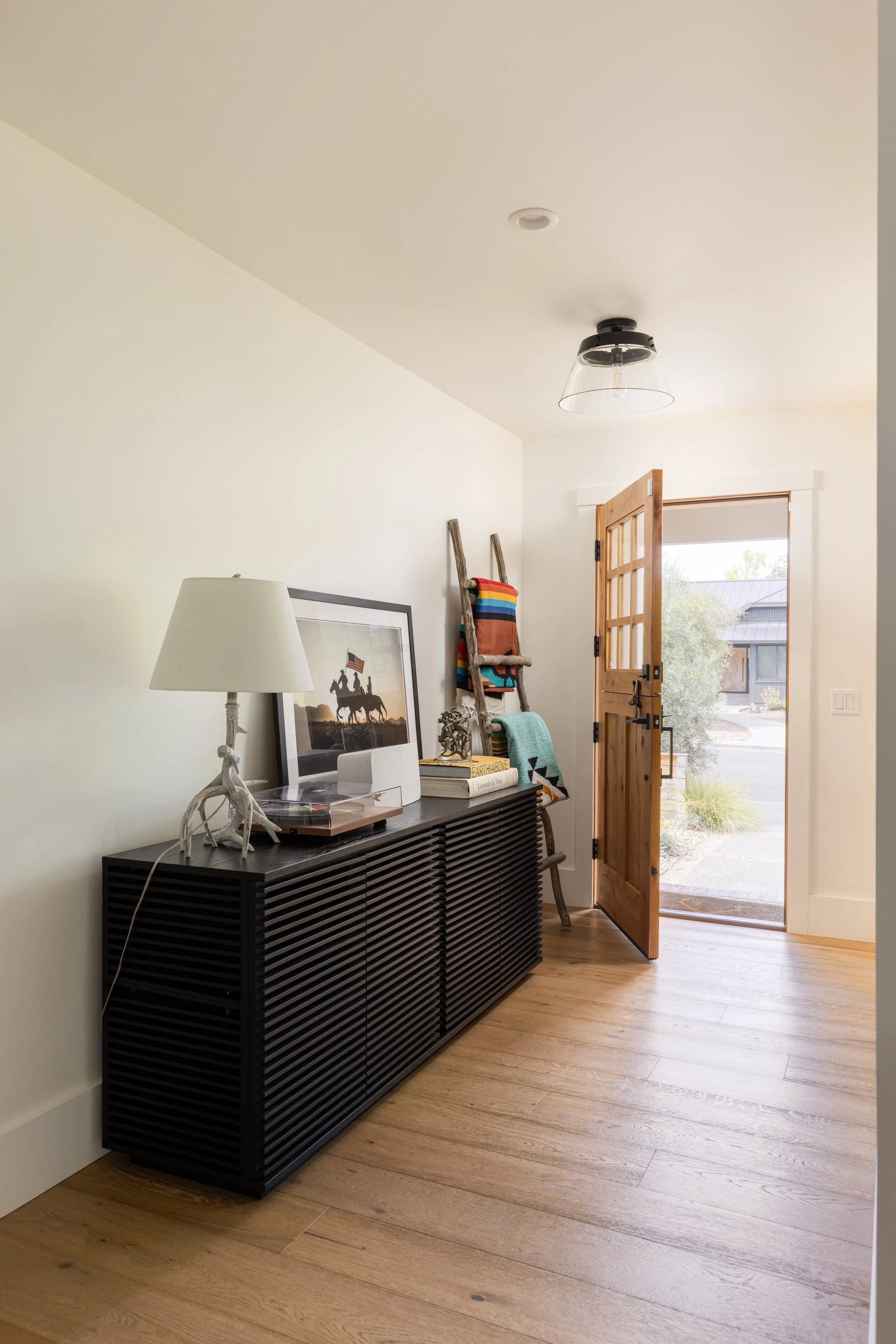 Interior of a home with a wooden front door open to the outside, a black sideboard with decorative items and a lamp, and a ladder with a colorful blanket leaning against the wall.