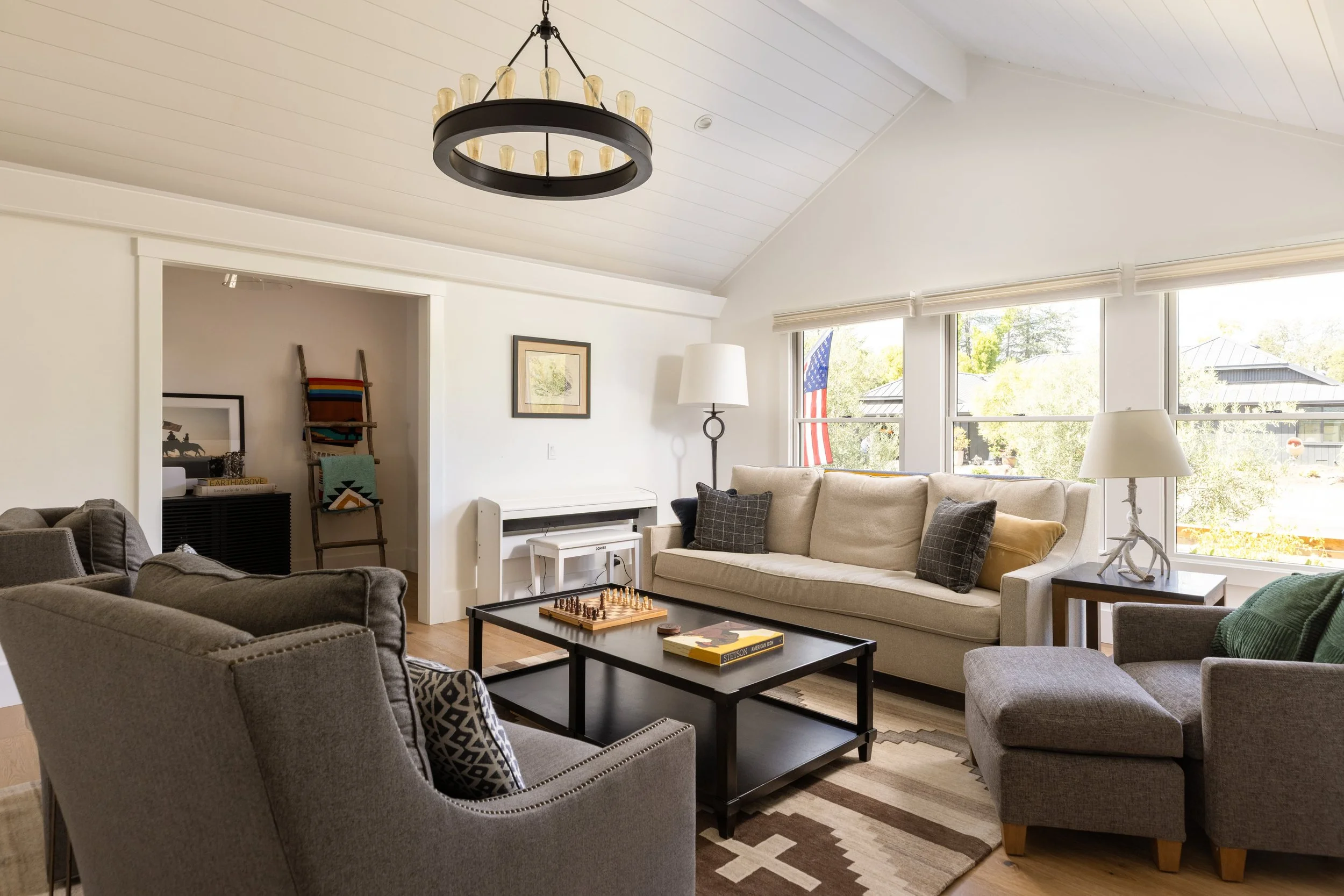 A cozy living room with beige sofas, colorful throw pillows, and a black coffee table with a chess set. Large windows with blinds reveal trees and a house outside. A white piano, framed artwork, and a ladder with colorful blankets are in the backgrou