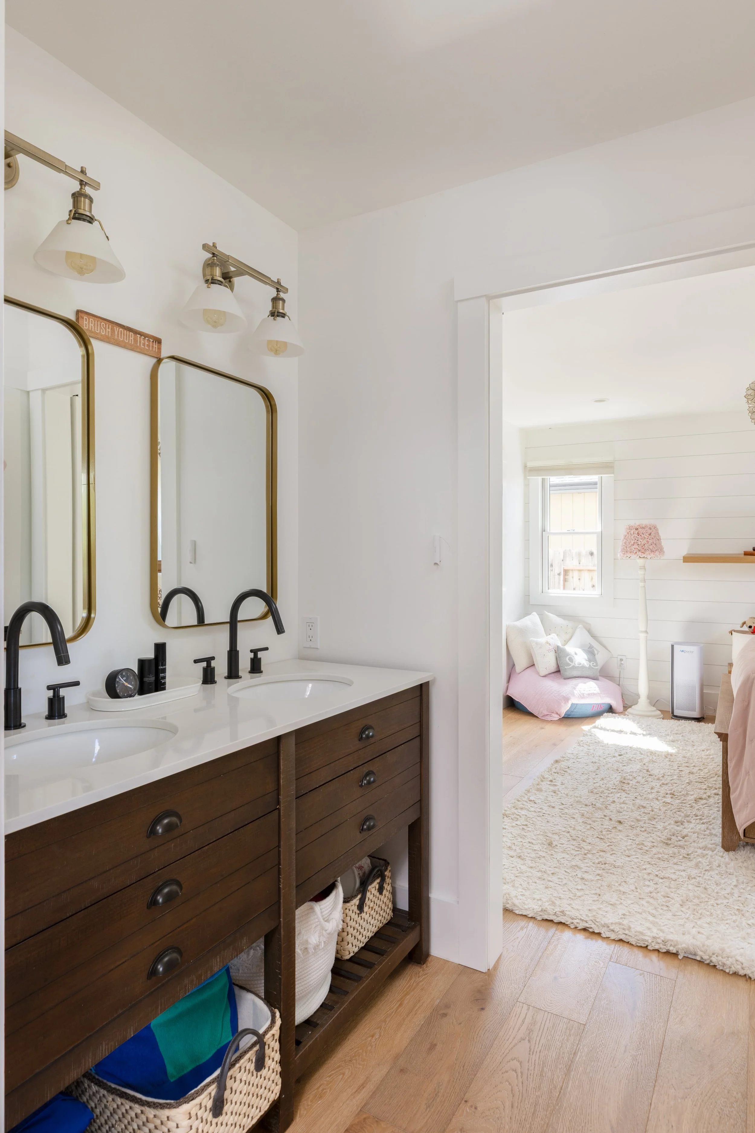 A bathroom with two sinks, dark wood vanity, three mirrors, and three wall-mounted light fixtures, leading into a light-filled bedroom with white walls, a window, pink bedding, a fluffy white rug, and decorative items.