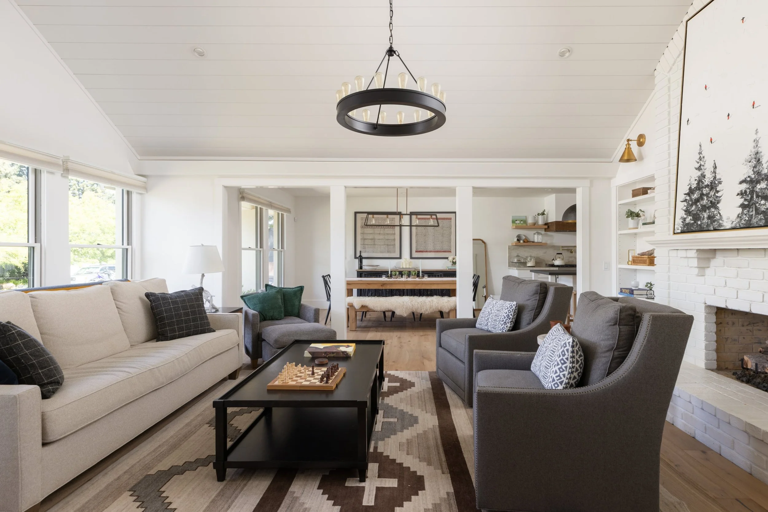 Living room with beige and gray sofas, black coffee table with chess set, patterned rug, white brick fireplace, and large windows with blinds