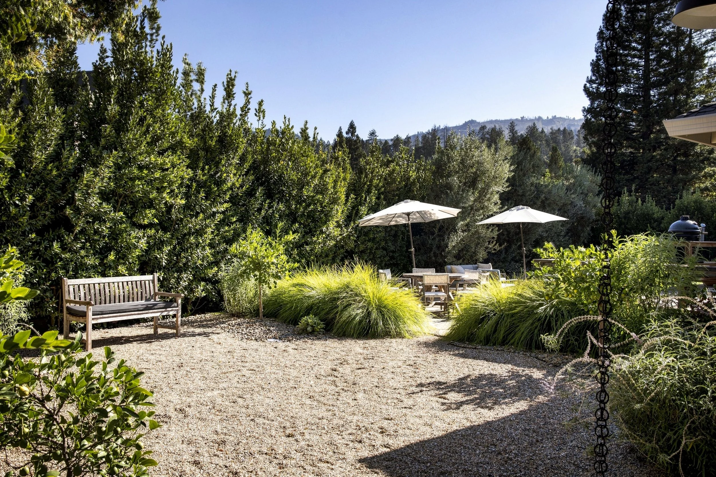 A garden with a gravel pathway, benches, white umbrellas, lush green shrubs, and tall trees with hills in the background under a clear sky.