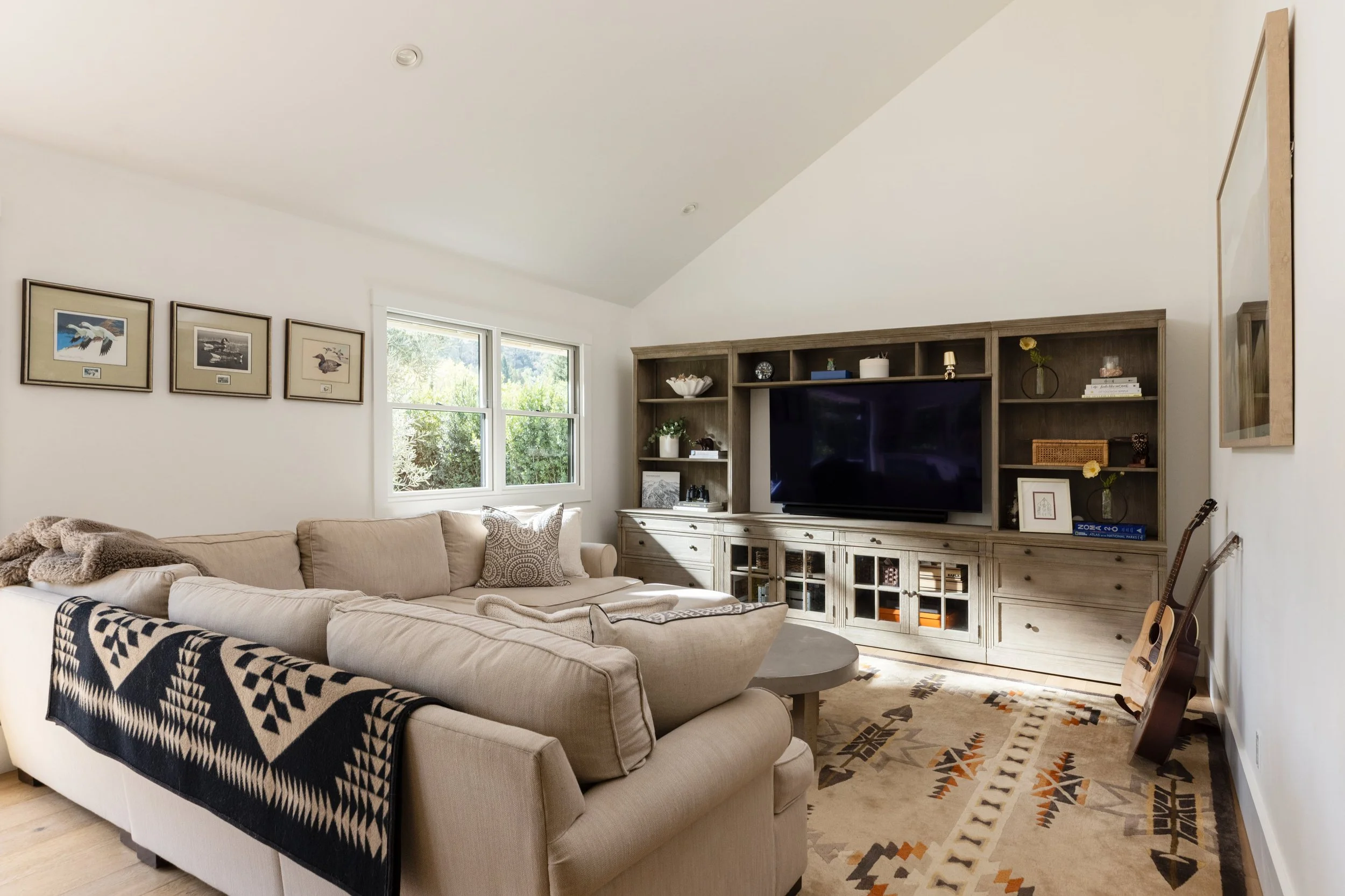 A cozy living room with a beige sectional sofa, black and white patterned blanket, and decorative pillows, set in front of a wooden entertainment center with a flat-screen TV and various decor items, window with view of greenery, and a colorful patte