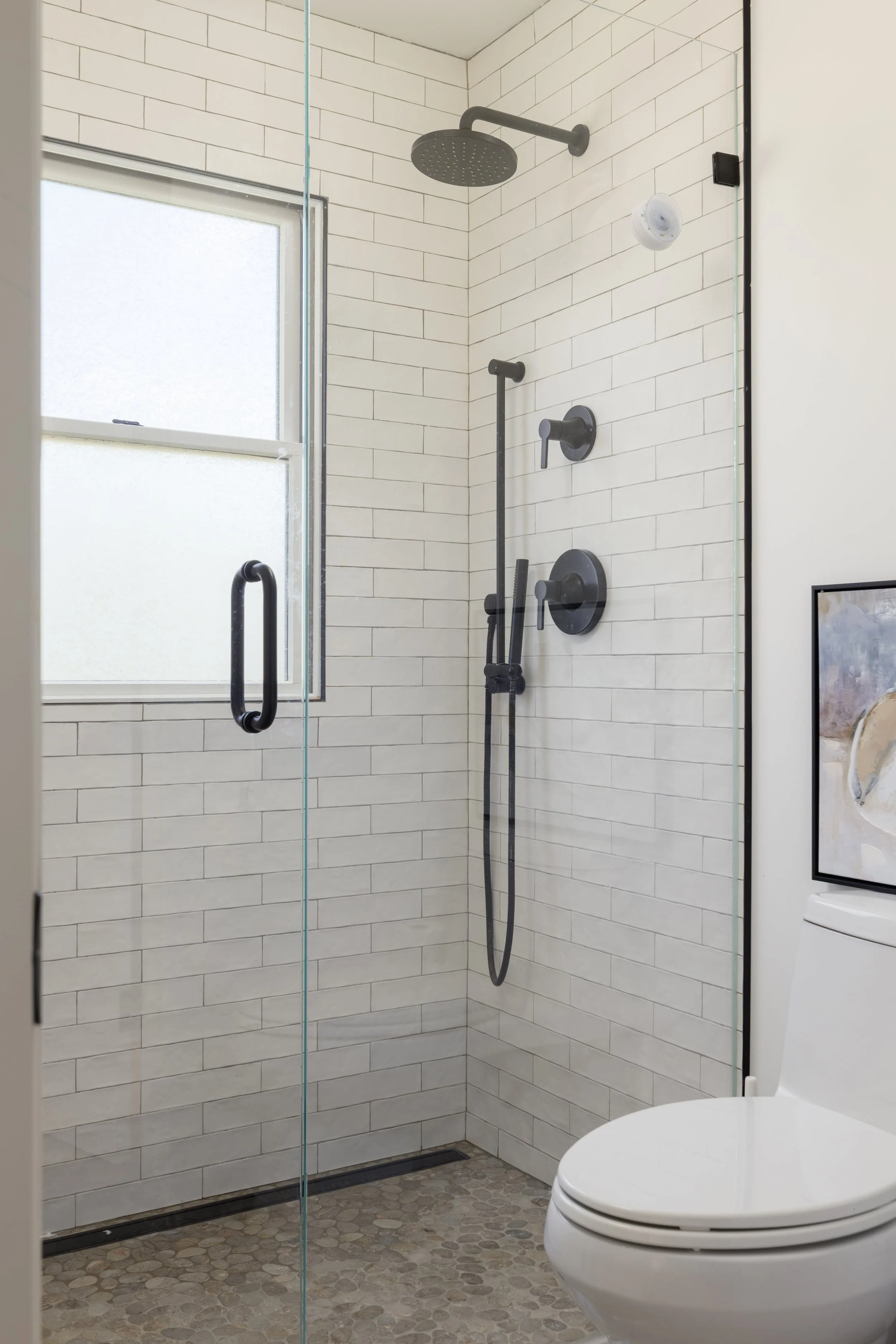 A modern bathroom shower area with white tile walls, a frosted window, a black shower head, a handheld shower, and a toilet to the right.