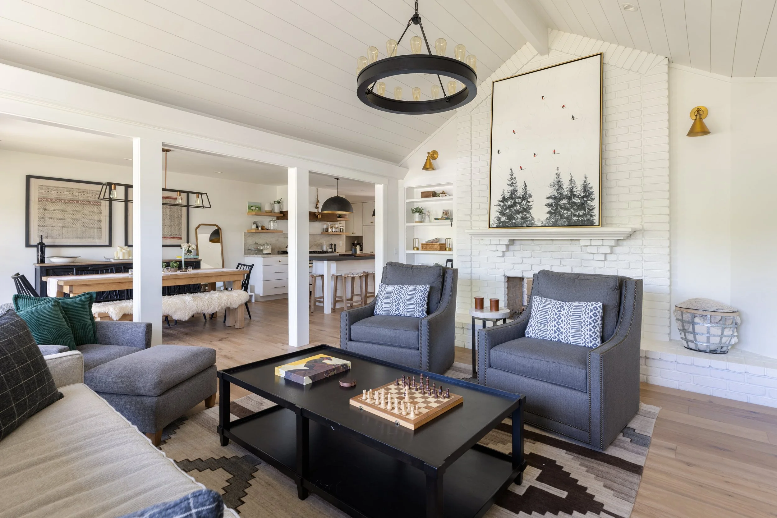 A living room with a black coffee table featuring chess and dominoes, gray armchairs, a beige sofa, and patterned throw pillows. The room has a white brick fireplace, large wall art depicting trees, and modern wall-mounted gold lights. The background
