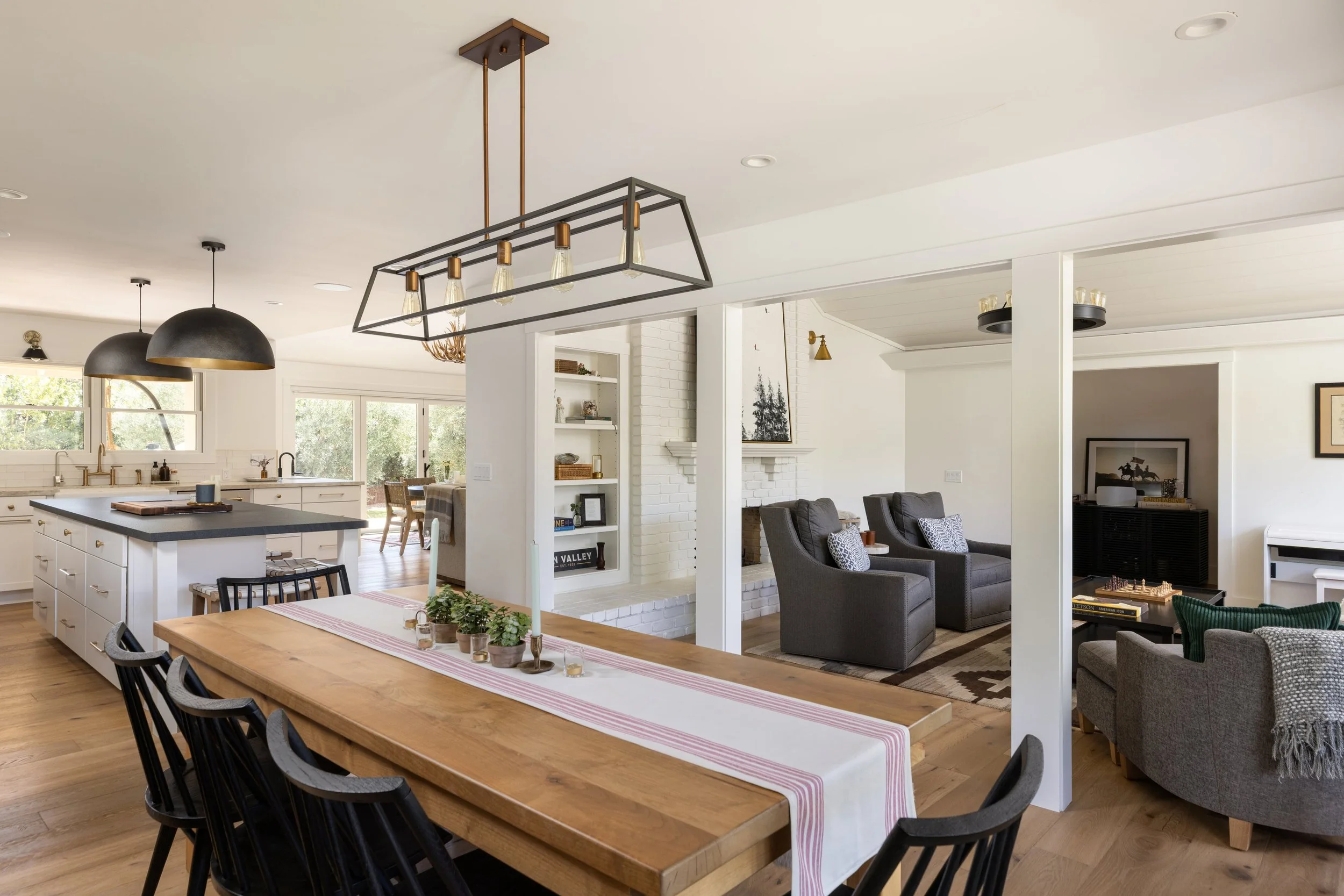 Open-concept kitchen and living room with modern lighting, white cabinets, a large wooden dining table, and gray seating area.