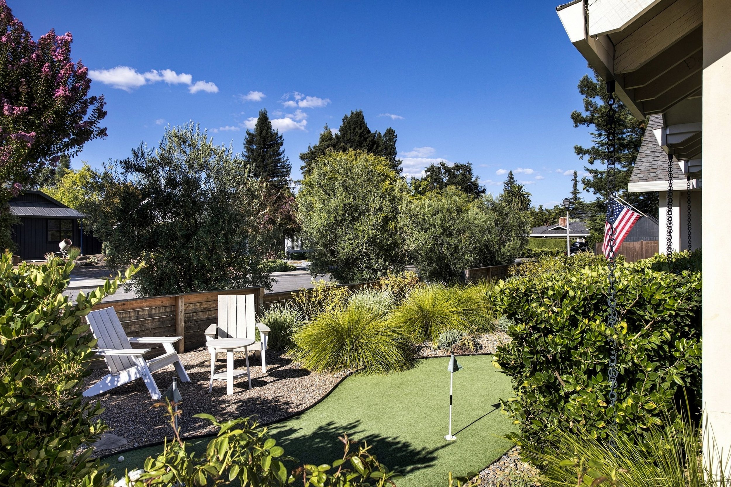A backyard with Adirondack chairs and a putting green, surrounded by greenery and trees, with a house to the right and a blue sky with scattered clouds above.