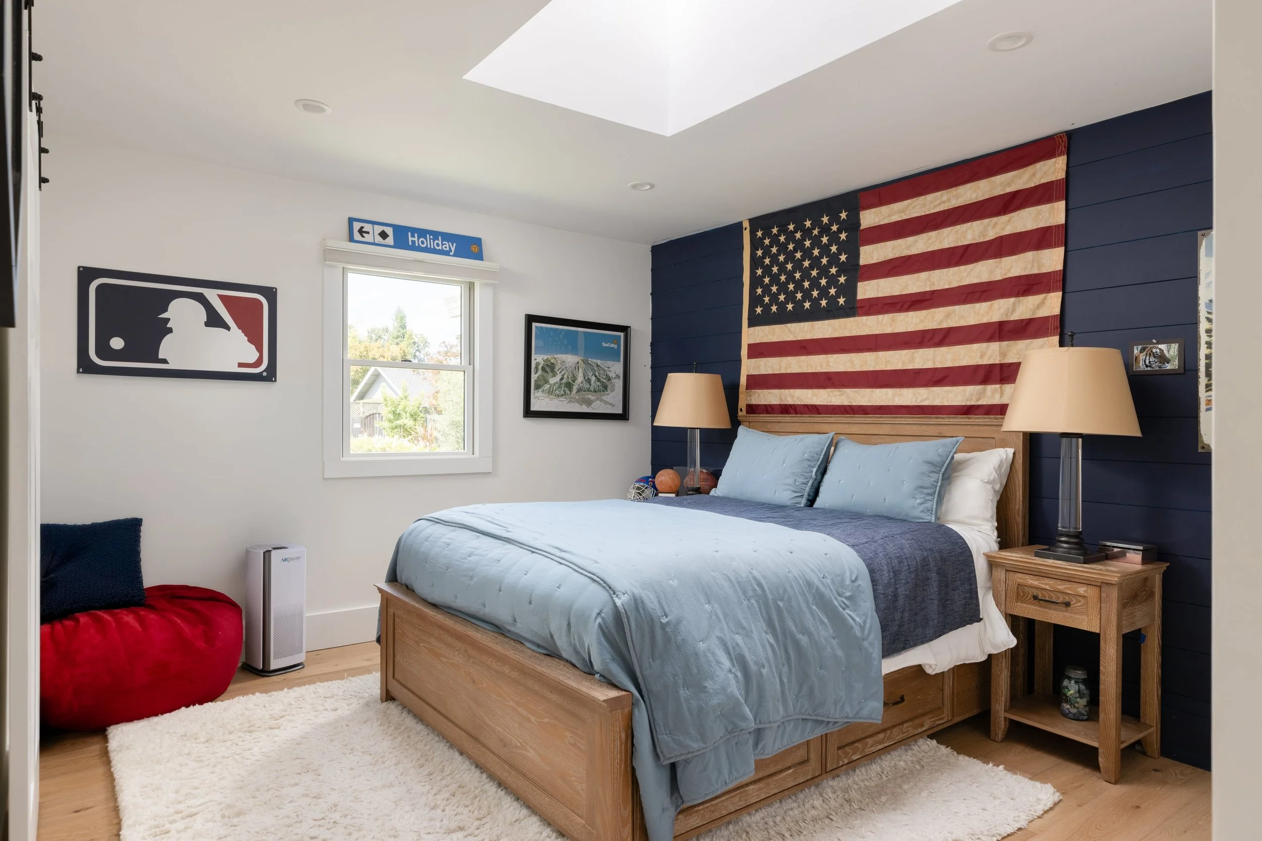 A bedroom with a wooden bed, blue bedding, and an American flag hanging on a navy blue side wall. There are two beige lamps on wooden nightstands, artwork on the walls, a red bean bag, and a white furry rug on a wooden floor.