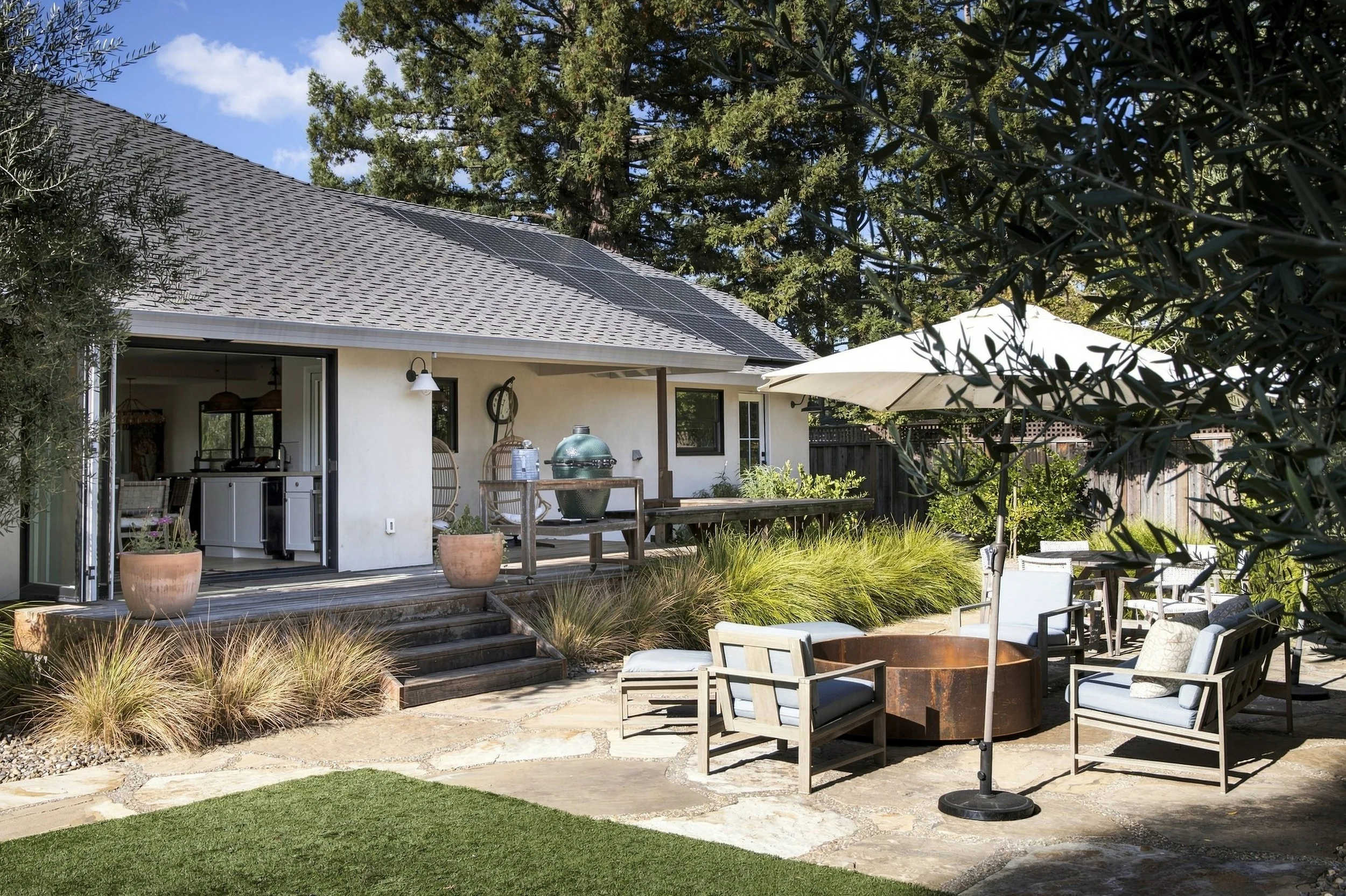 Backyard patio with outdoor furniture, potted plants, and a house with solar panels on the roof, surrounded by trees and greenery.