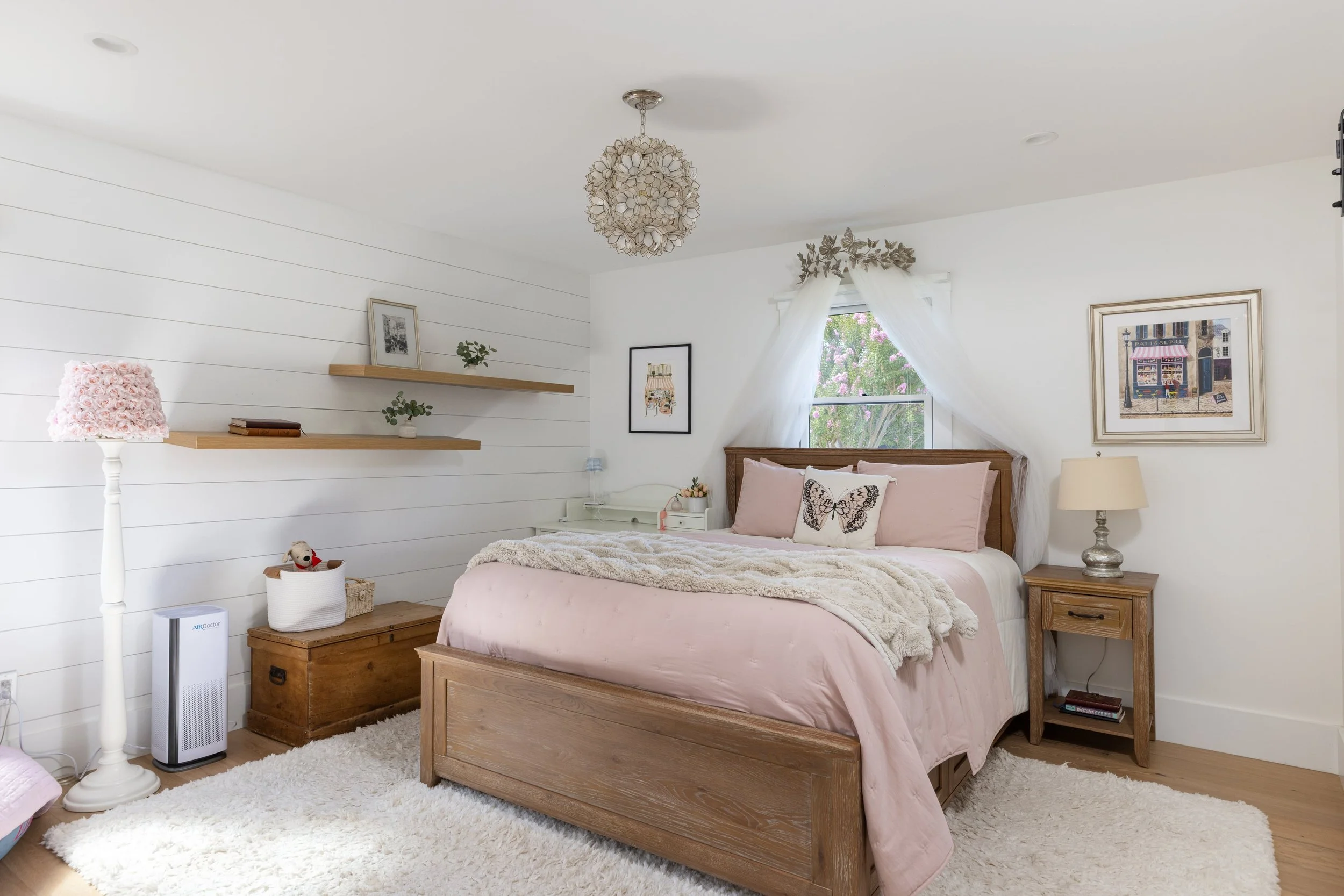 A cozy bedroom with a wooden bed frame and pink bedding, white walls with shiplap, a window with white curtains, and various framed pictures. The room has a white fluffy rug and a bedside table with a lamp. There is a white lampshade on a stand, a sm