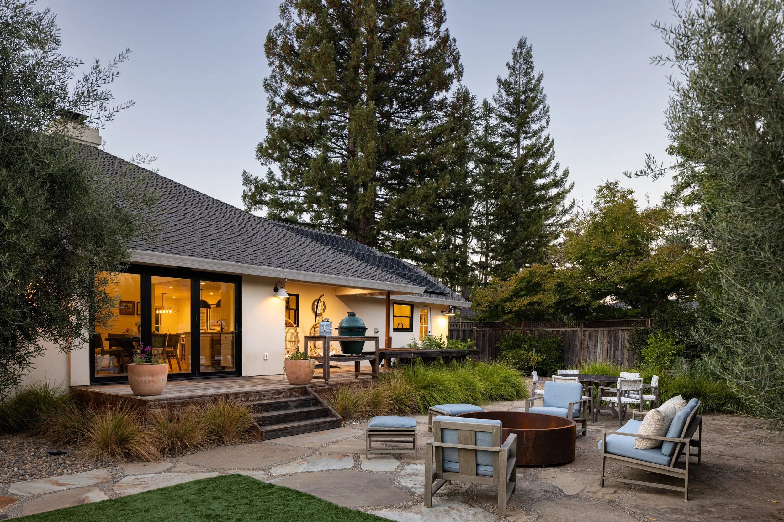 A backyard patio area during dusk with outdoor furniture and potted plants, adjacent to a modern house with large glass sliding doors, surrounded by trees and a wooden fence.