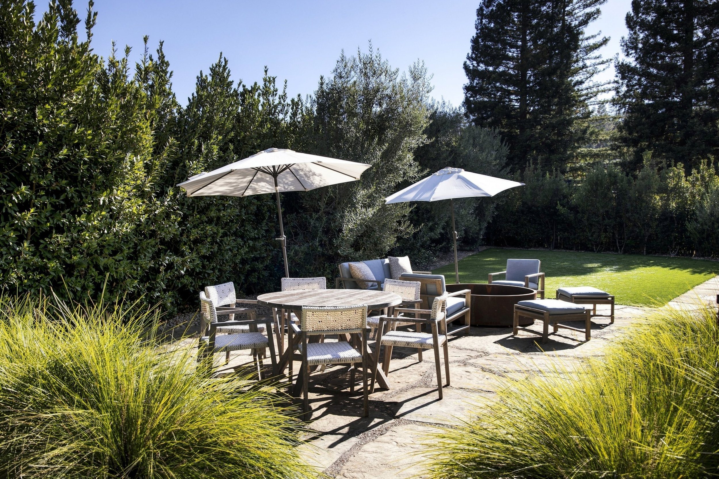 An outdoor patio area with white umbrellas, a round wooden table, and several chairs surrounded by lush green plants and trees under a clear blue sky.