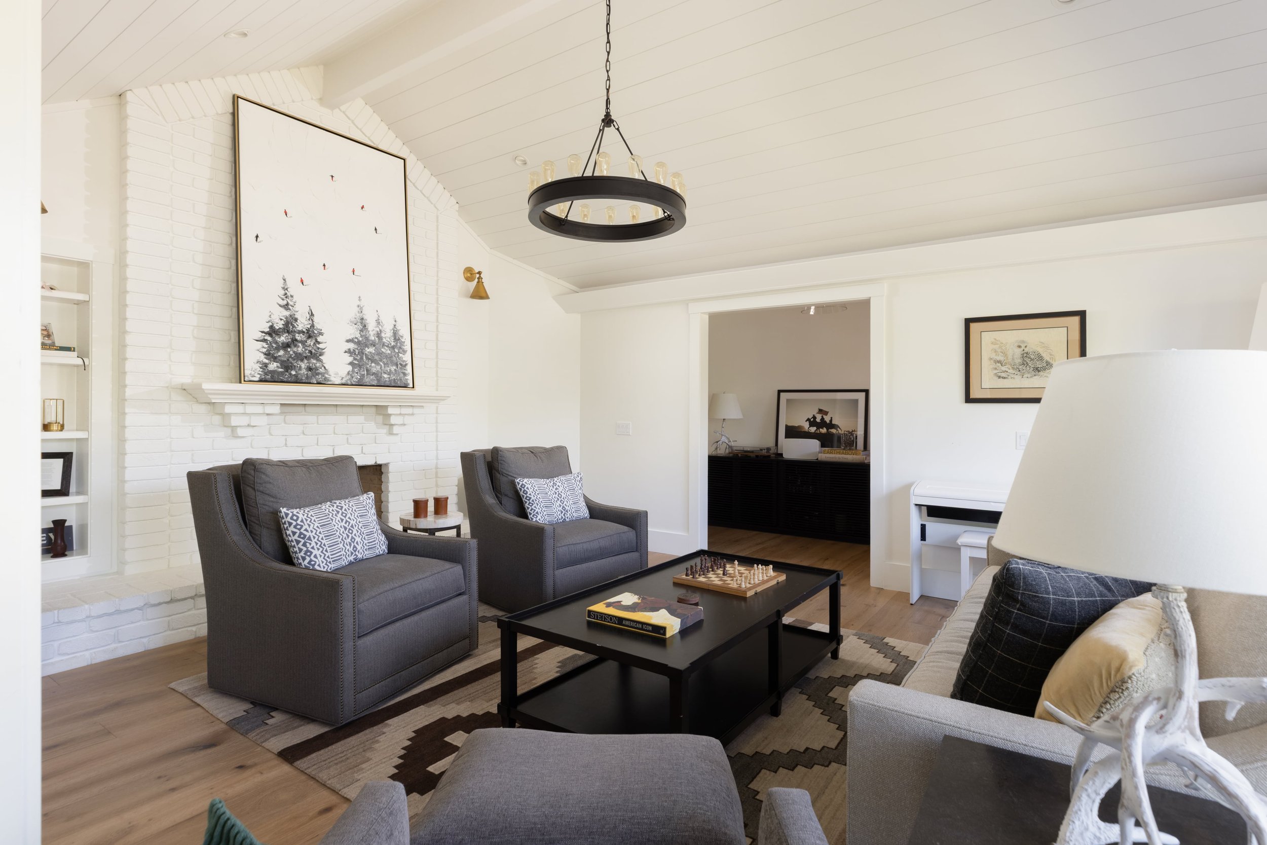 A living room with white painted brick fireplace, two grey armchairs with patterned pillows, a black coffee table with a chess set and books, a white piano, framed artwork on the walls, a modern chandelier, and hardwood flooring.