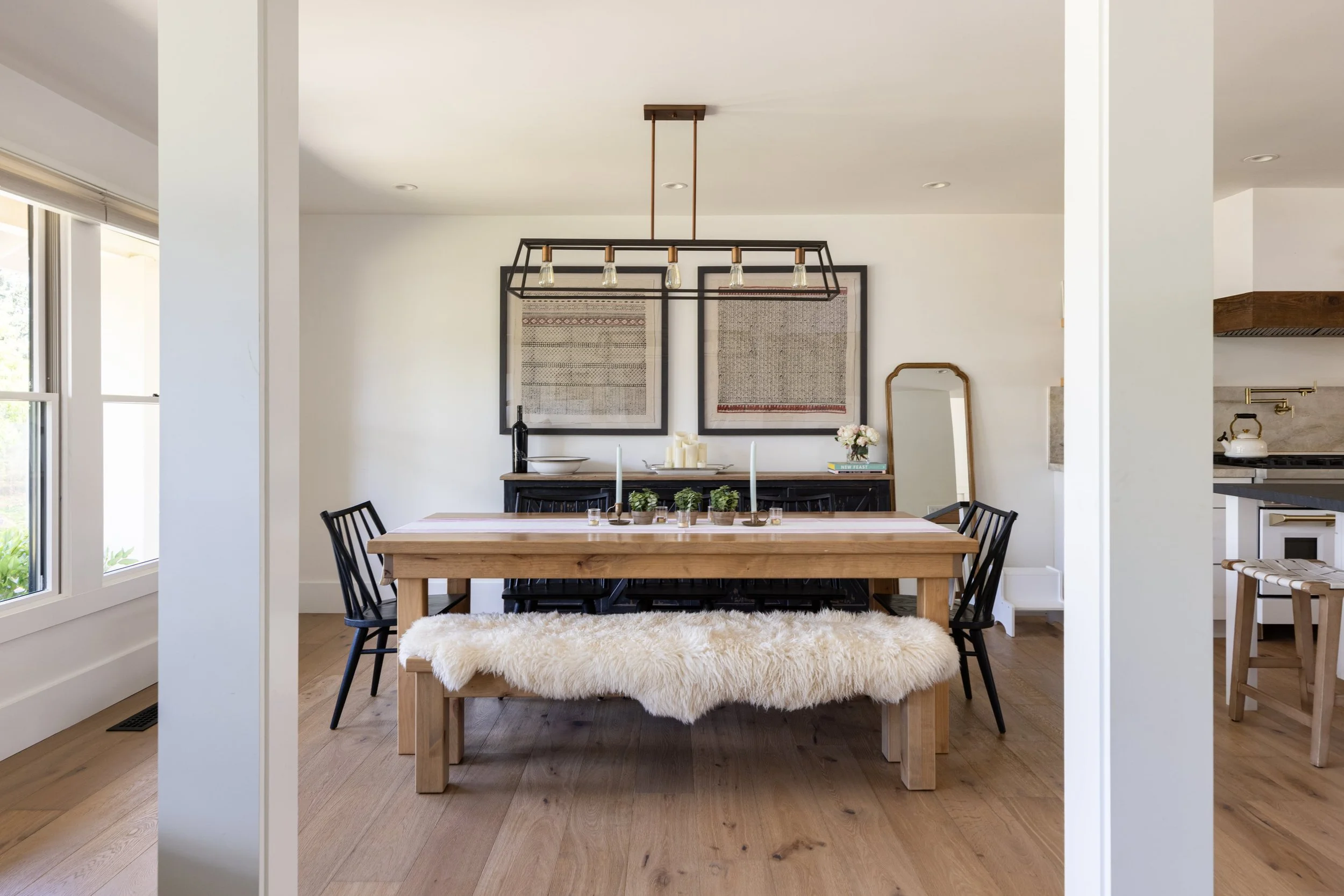 A dining room with a wooden table, black chairs, and a fluffy white bench. The wall features two framed textile art pieces, with candles, a wine bottle, a bowl, and flowers on a sideboard. A large window on the left provides natural light.