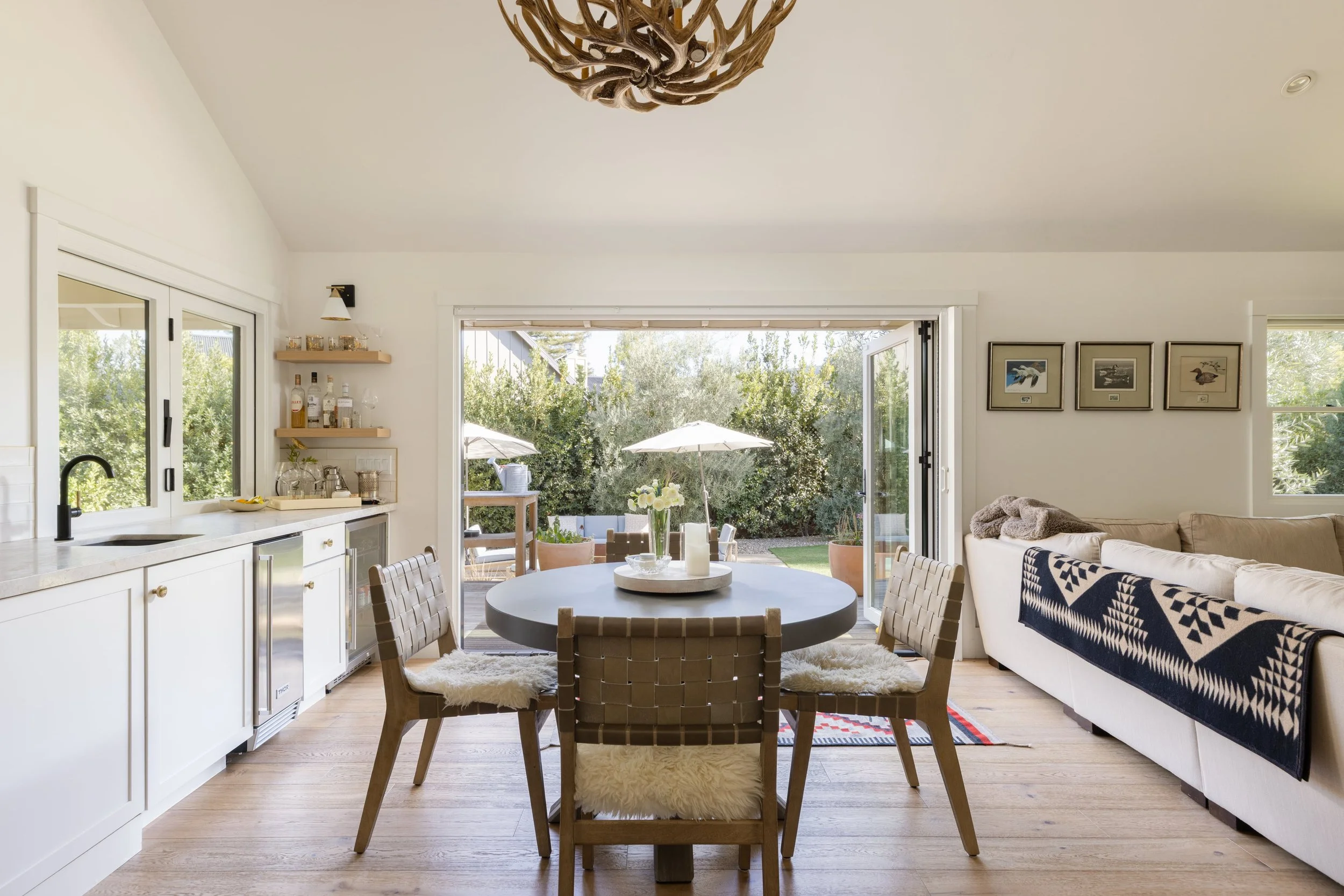 Bright, open living and dining area with large glass doors leading to a backyard patio with umbrellas and outdoor furniture. The room features a round dining table with woven chairs, a white sofa with patterned cushions, framed wall art, and a ceilin