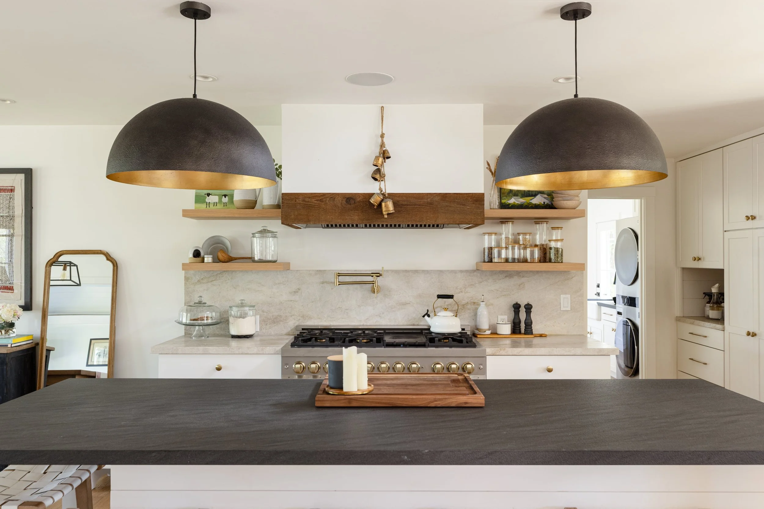 Modern kitchen with black pendant lights, white cabinets, a marble backsplash, and a stove. Decor includes glass jars, dishes, a mirror, and candles.