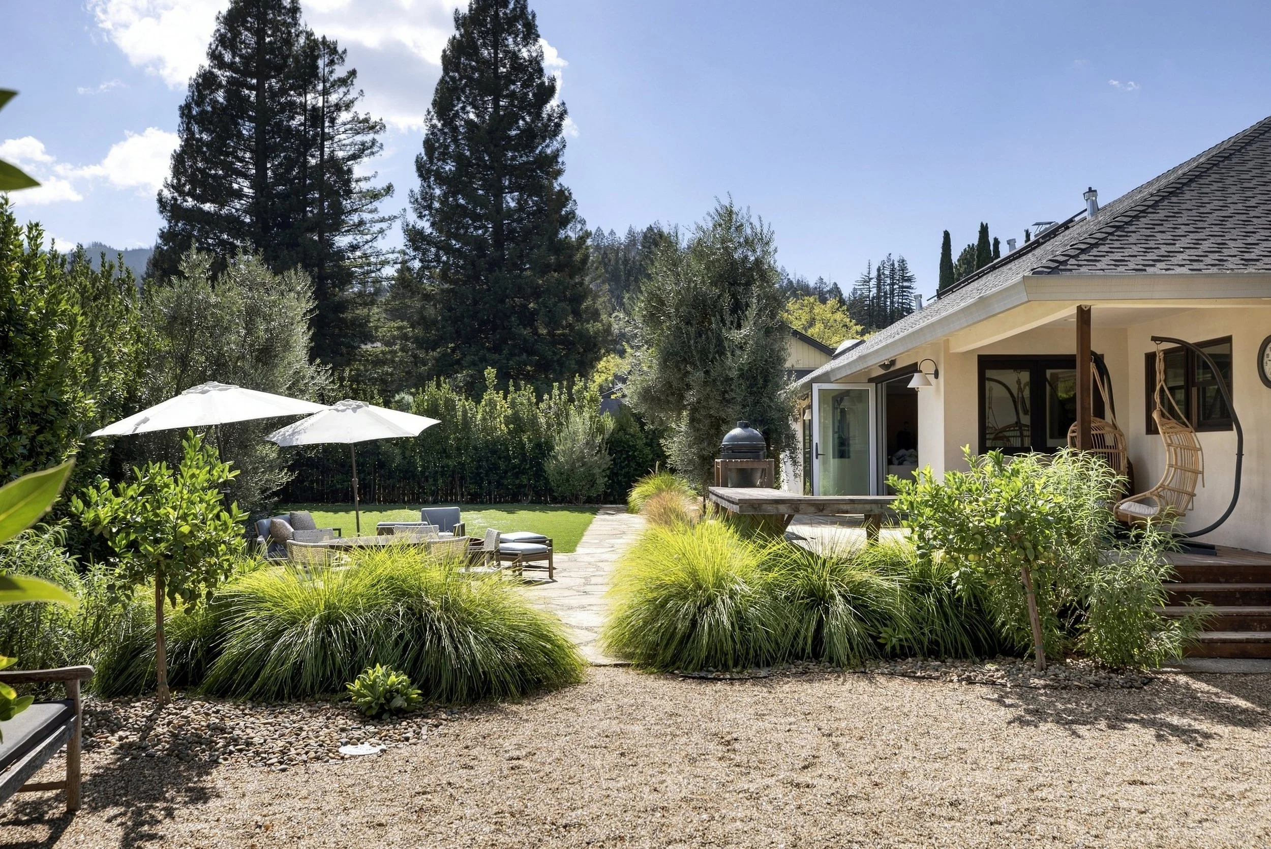 A backyard with a gravel path, outdoor furniture, umbrellas, and lush green plants. A house with a patio and a wooded area with tall trees in the background.