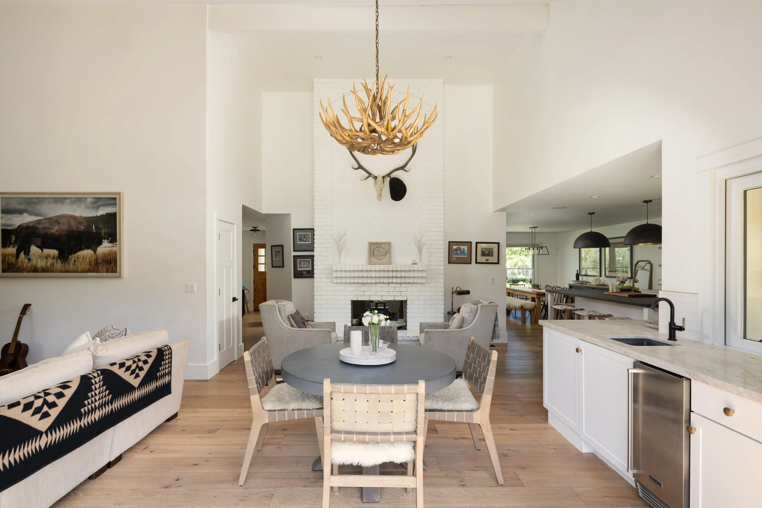 Spacious living room with white walls, a white brick fireplace with antler chandelier and mounted deer skull, surrounded by framed pictures, beige armchairs, and a white sofa with black geometric throw, wooden accents, and a round table with a vase o