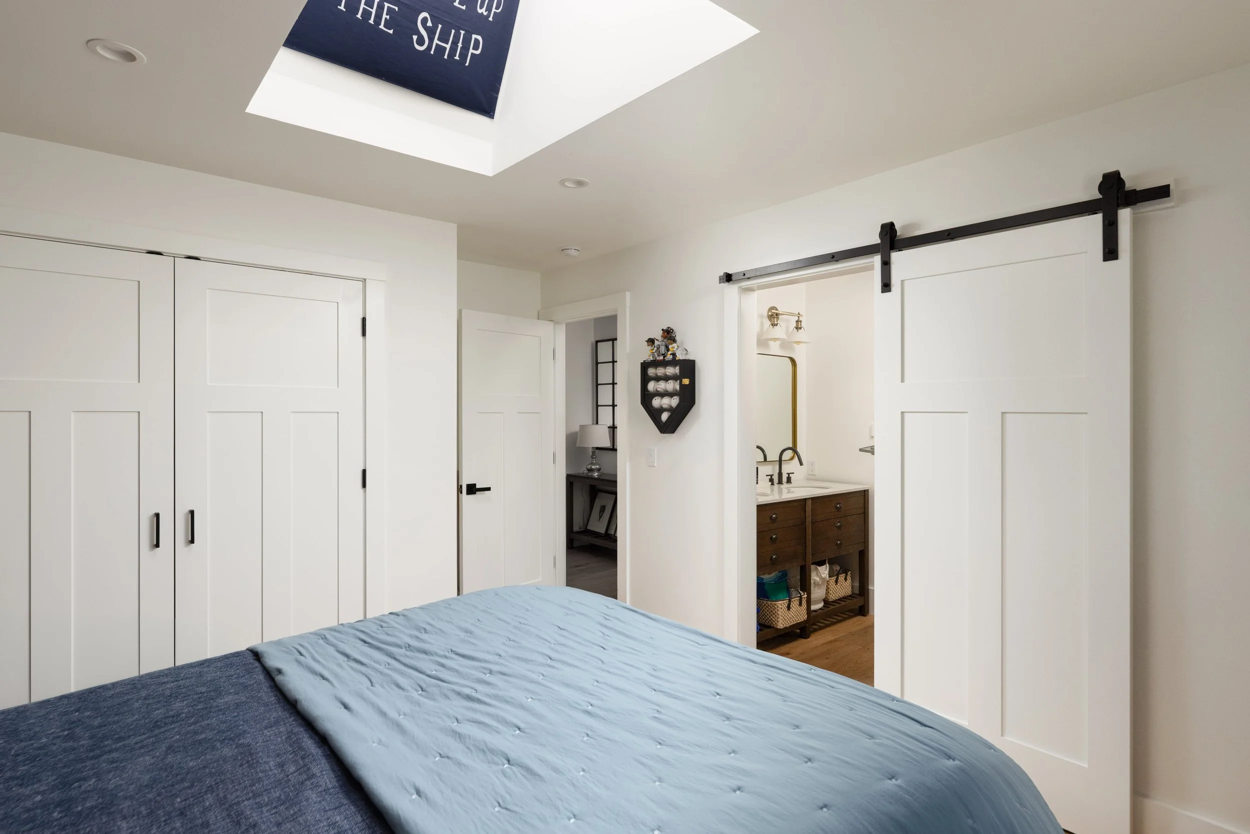 A bedroom with a partially made bed with a blue quilted mattress cover. White sliding closet doors and a doorway leading to an additional room with a small black bookshelf displaying figurines, a lamp, and other decor. The ceiling has a skylight with