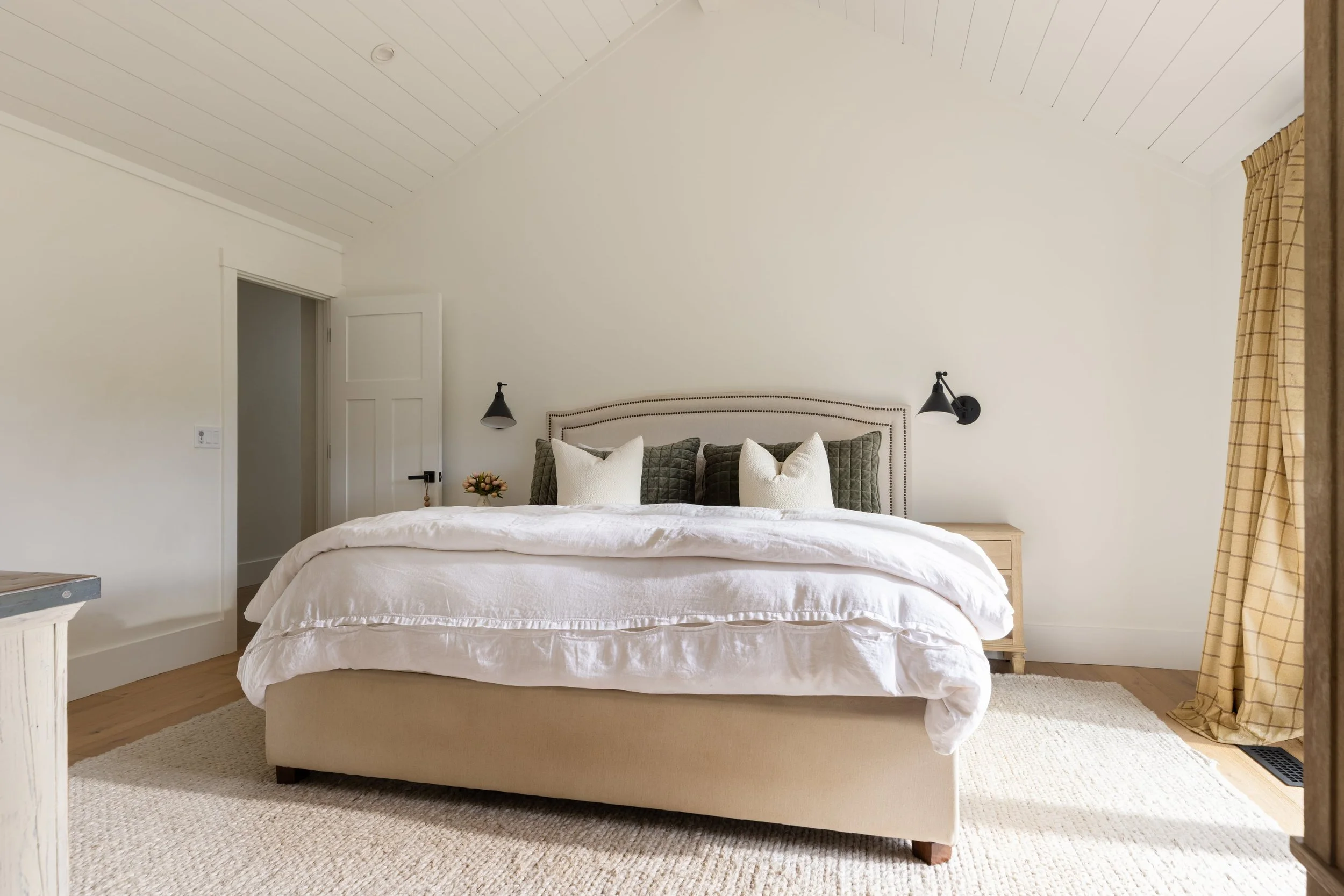 A neatly made bed with a beige upholstered bed frame, white bedding, and a variety of pillows against a white wall. There are black wall-mounted reading lamps on each side of the bed, a small flower arrangement on the bedside table, and beige curtain