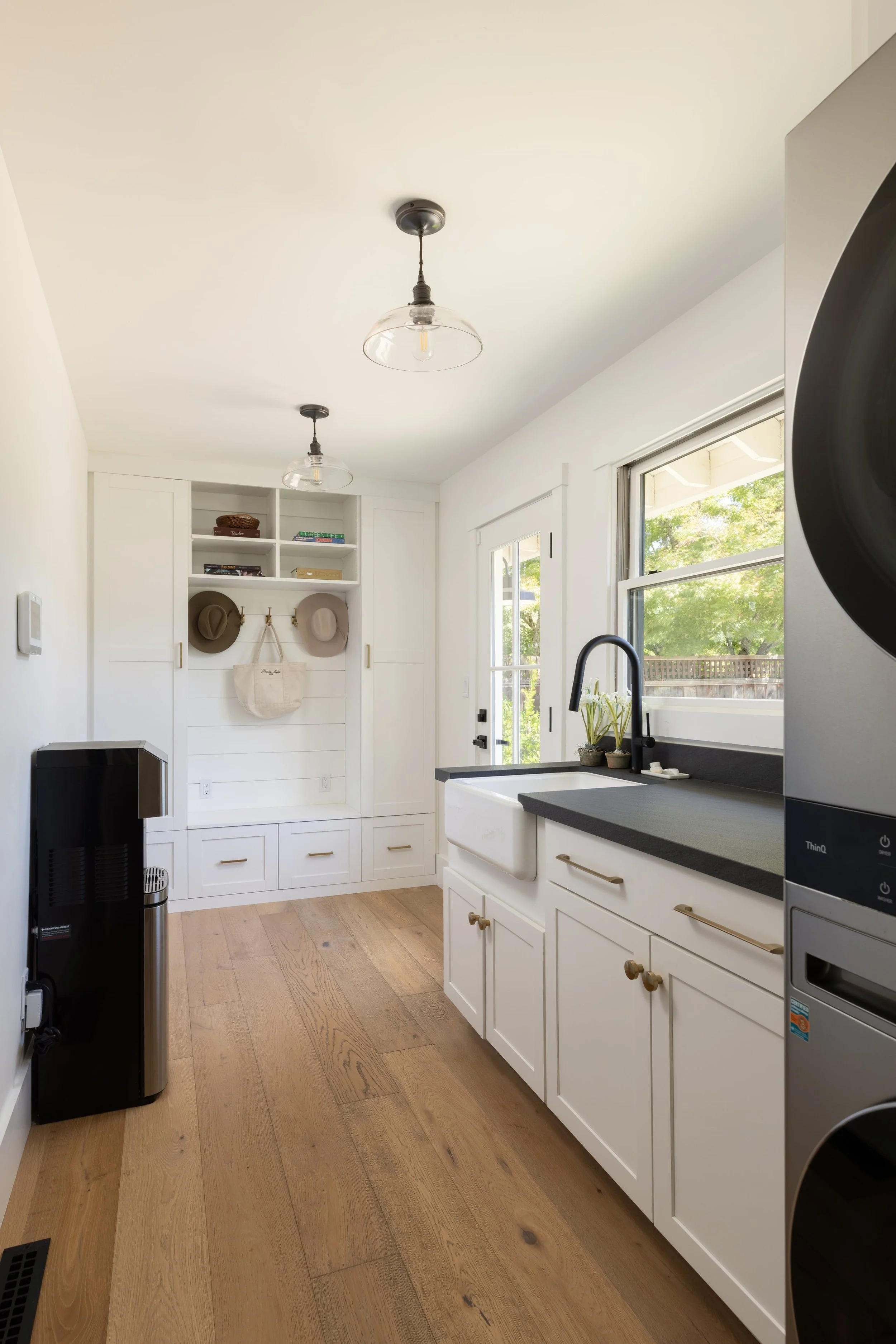 A modern kitchen with white cabinets, a black countertop, a farmhouse sink, a window overlooking a yard, and a wooden floor. Two ceiling light fixtures are also visible.
