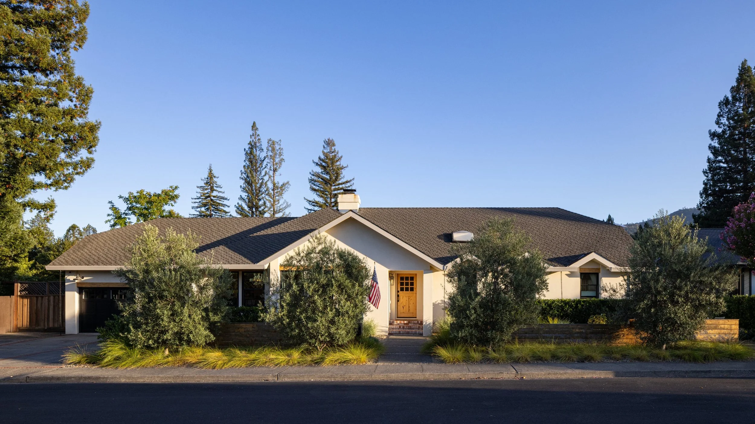 A house with a brown shingle roof, white walls, a small front porch, and an American flag by the entrance, surrounded by trees and shrubs under a clear blue sky.