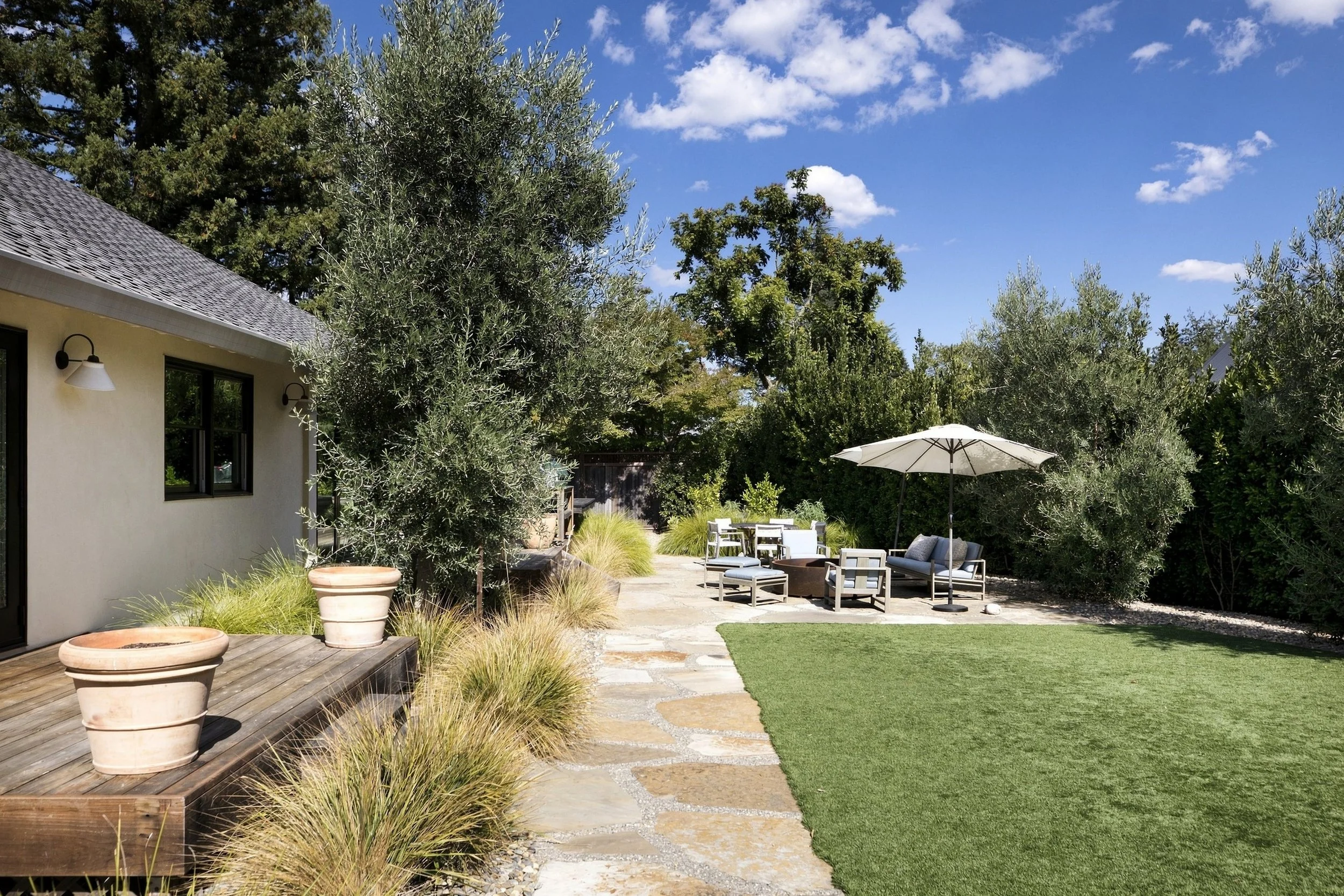 A backyard patio with outdoor furniture, umbrellas, large potted plants, and lush green trees under a blue sky with scattered clouds.