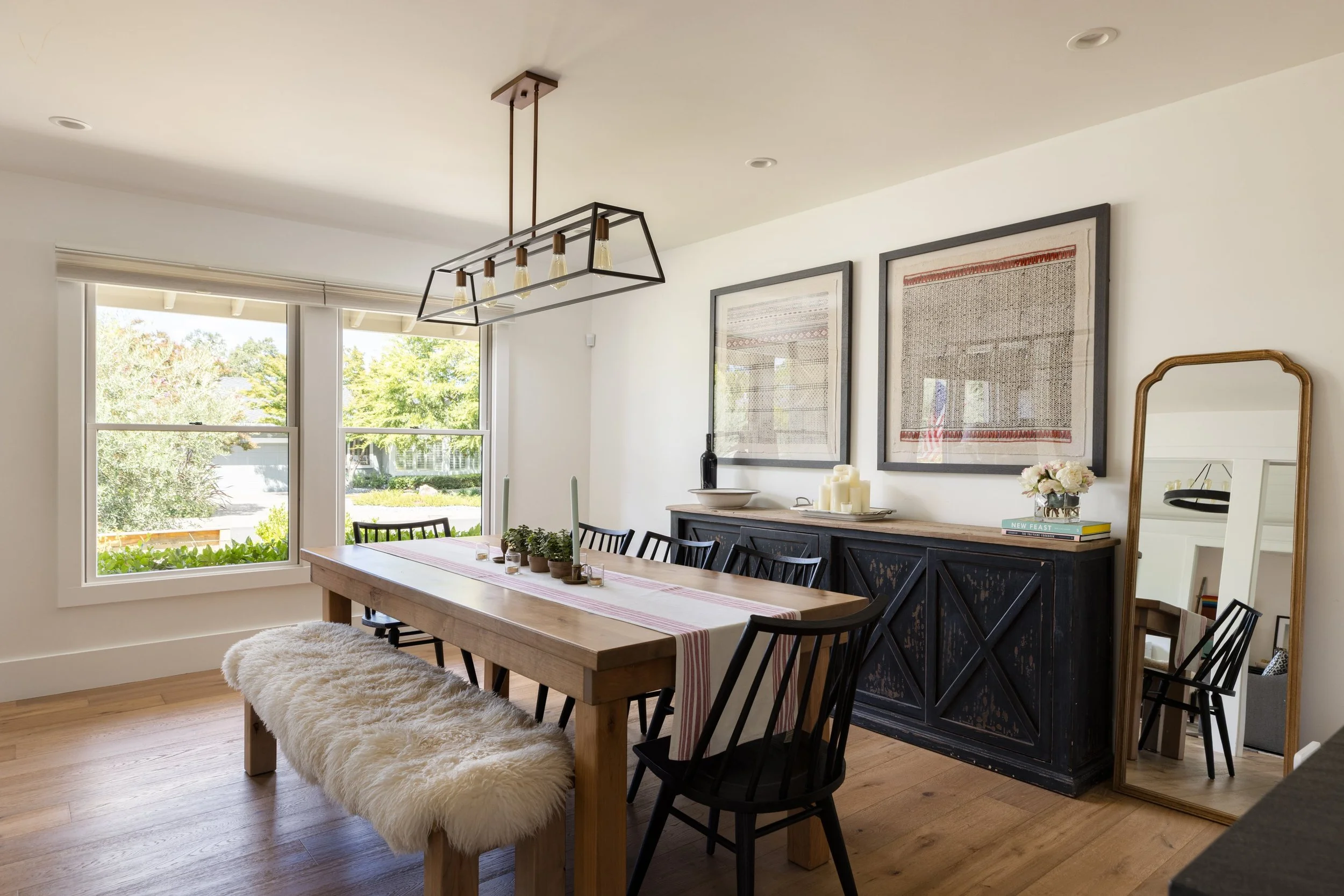 Dining room with a wooden table, black chairs, a fluffy white bench, wall art, a black sideboard with candles, and large windows with a view of trees.