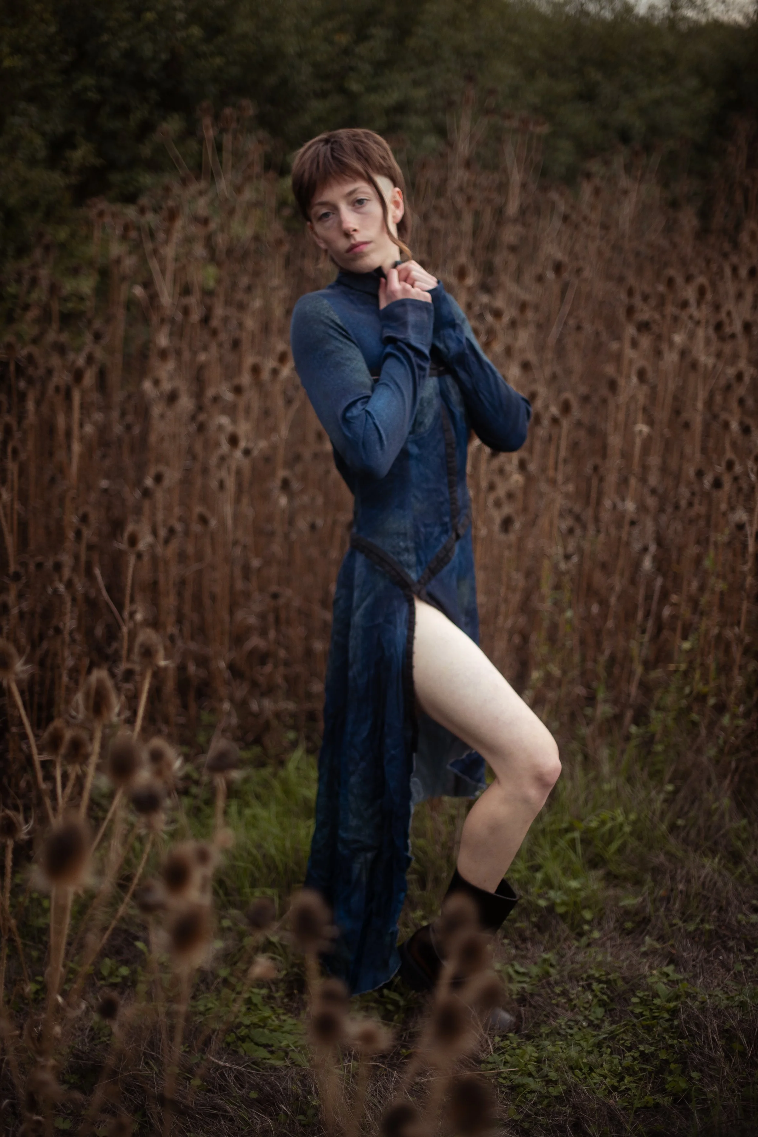 A person with short brown hair and pale skin wearing a blue long-sleeve garment standing outdoors in front of a dry, brown hedge, with one leg slightly raised and one hand near their neck.