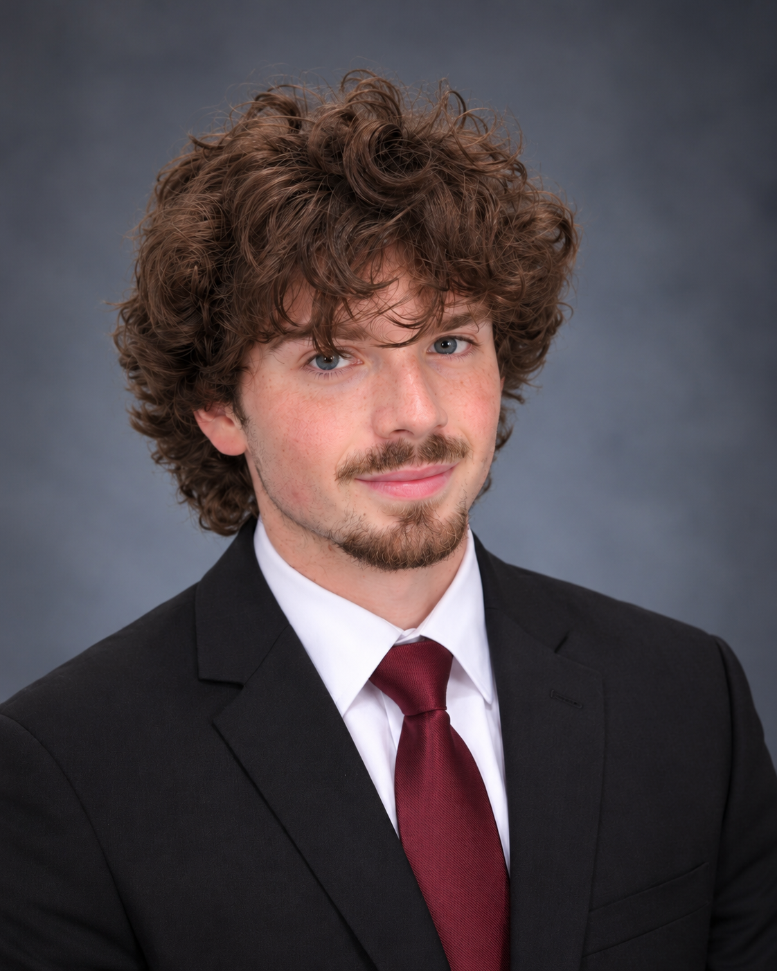 A young man with curly brown hair, blue eyes, and a light beard, wearing a black suit, white shirt, and burgundy tie, posing against a gray background.