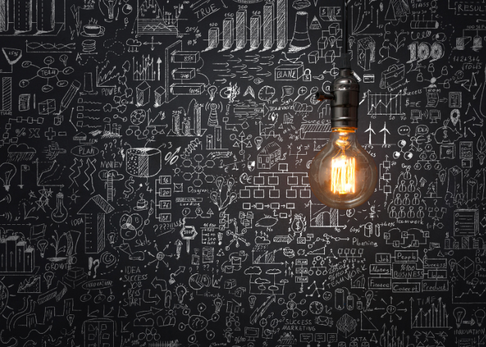 A glowing Edison-style light bulb hanging against a black wall filled with white chalk doodles of business concepts, charts, graphs, and ideas.