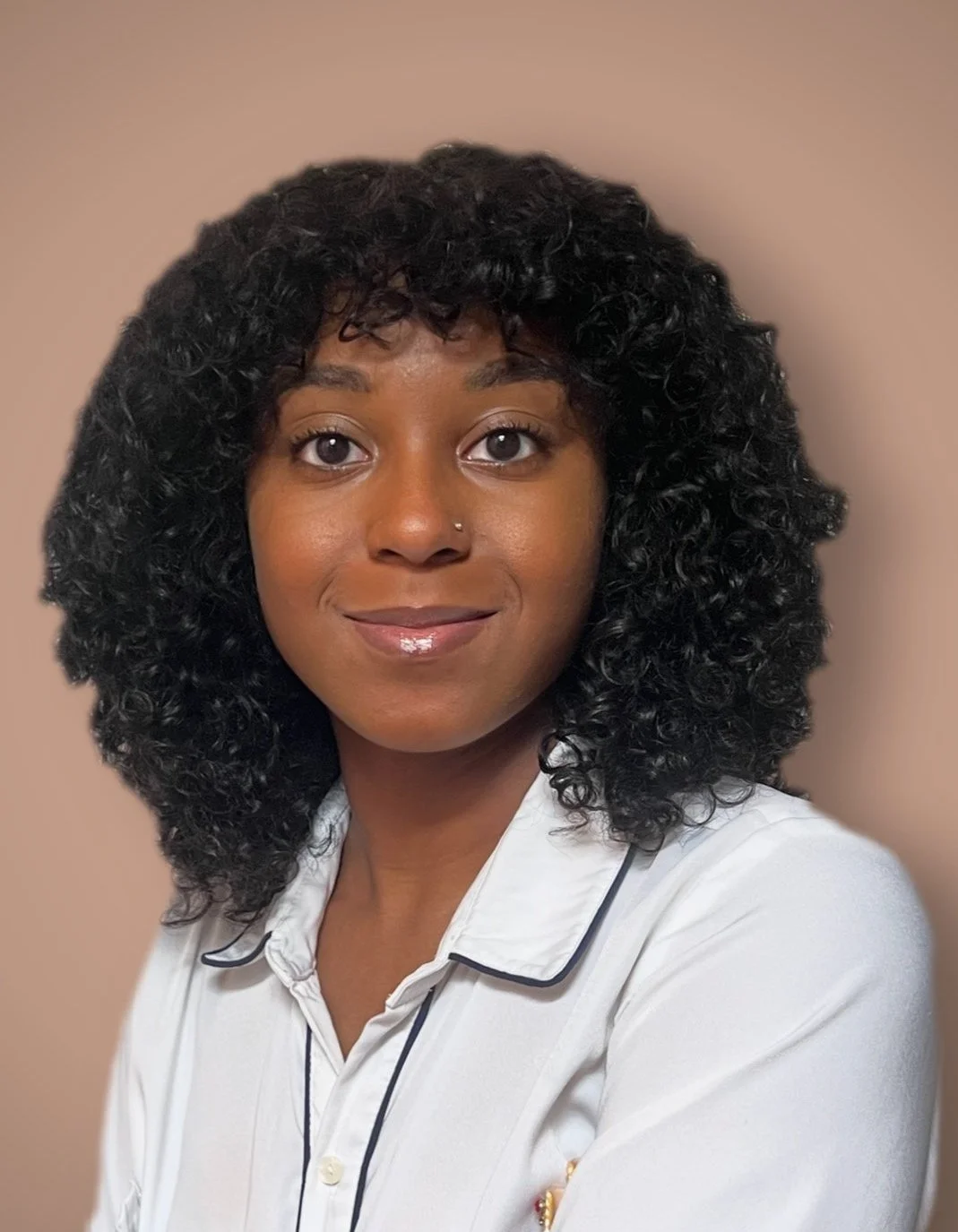 A woman with dark, curly hair and a nose piercing smiling at the camera. She is wearing a white shirt with black piping and a beige background.