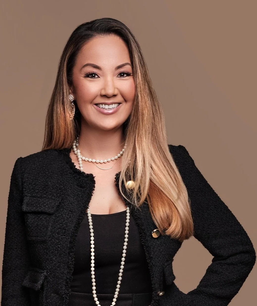 A woman with long, light brown hair and a friendly smile, dressed in a black top and textured black jacket, wearing pearl earrings, a pearl necklace, and layered jewelry, standing against a plain brown background.