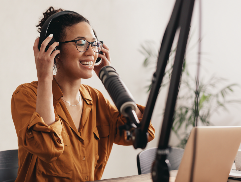 Woman with curly hair and glasses smiling while speaking into a microphone during a recording session, wearing a brown shirt, headphones on, with a laptop in front of her.