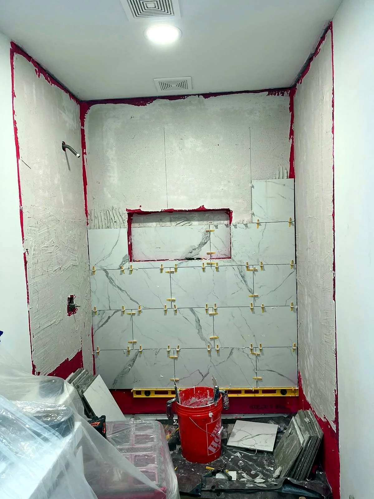 Bathroom under renovation with marble-look wall tiles, tile spacers, construction tools, and a red bucket