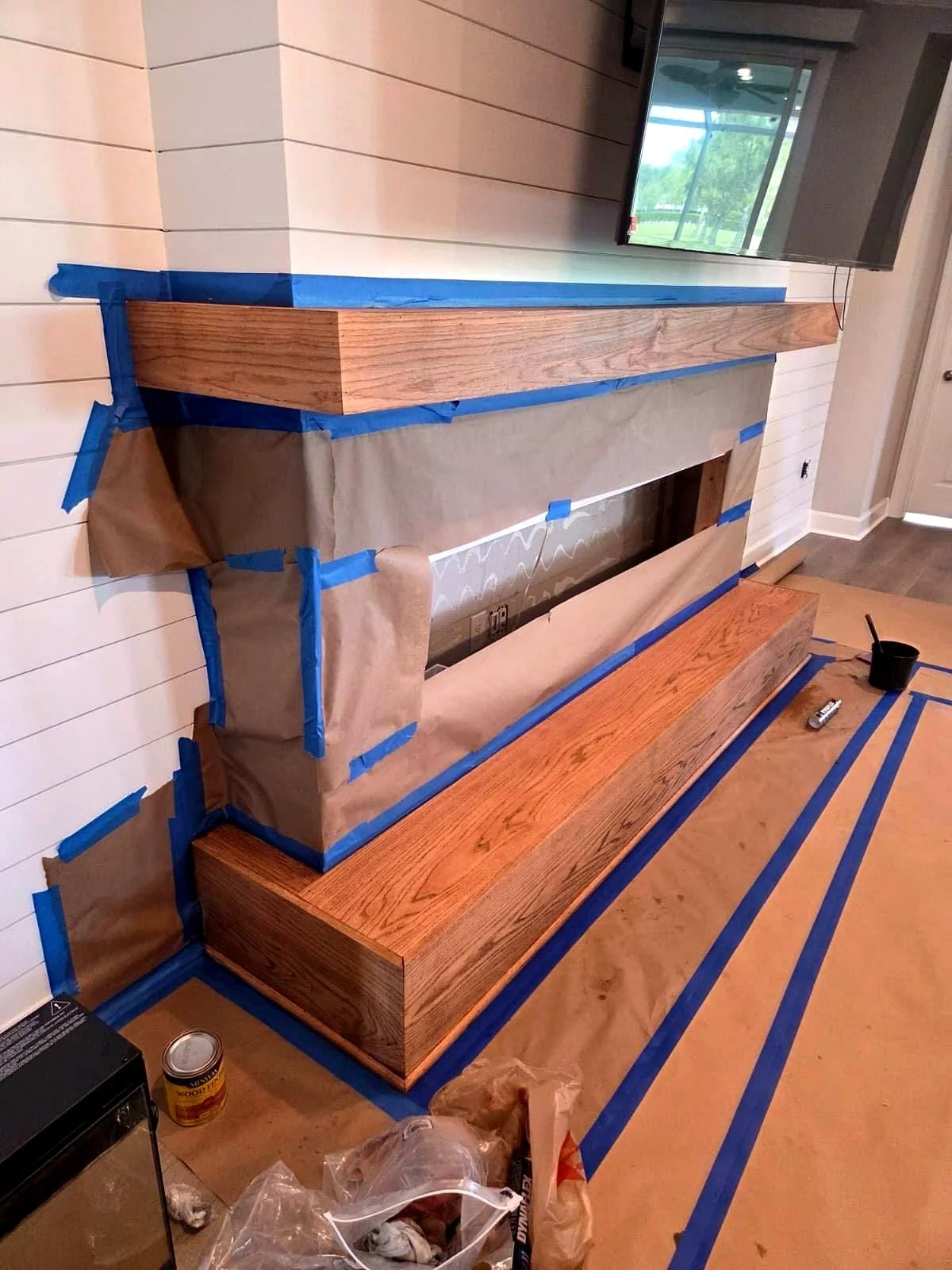 Living room fireplace under renovation, masked with brown paper and blue painter’s tape, with unfinished wood mantel and hearth.