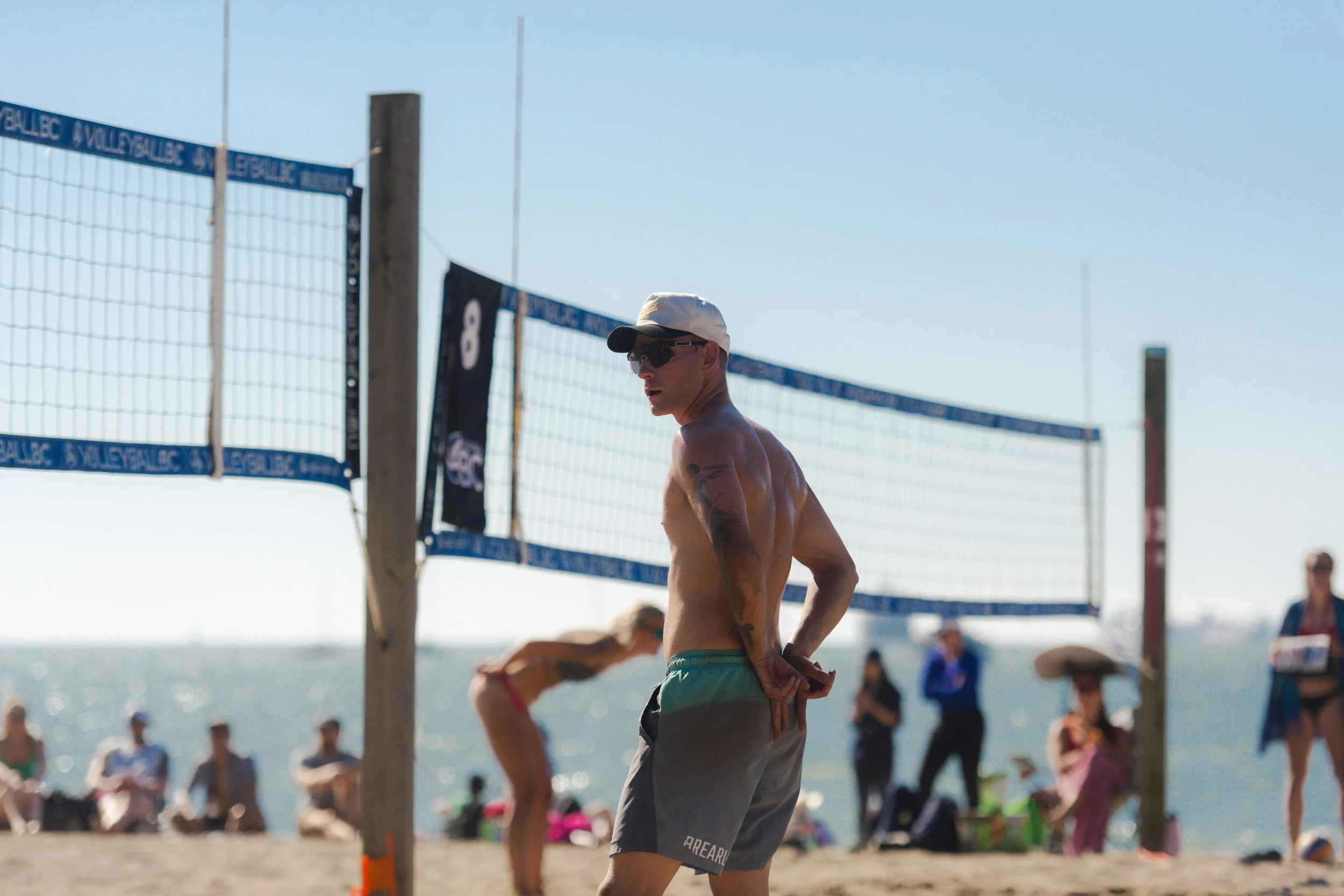 Shirtless beach volleyball player wearing a cap and sunglasses on a sunny beach court