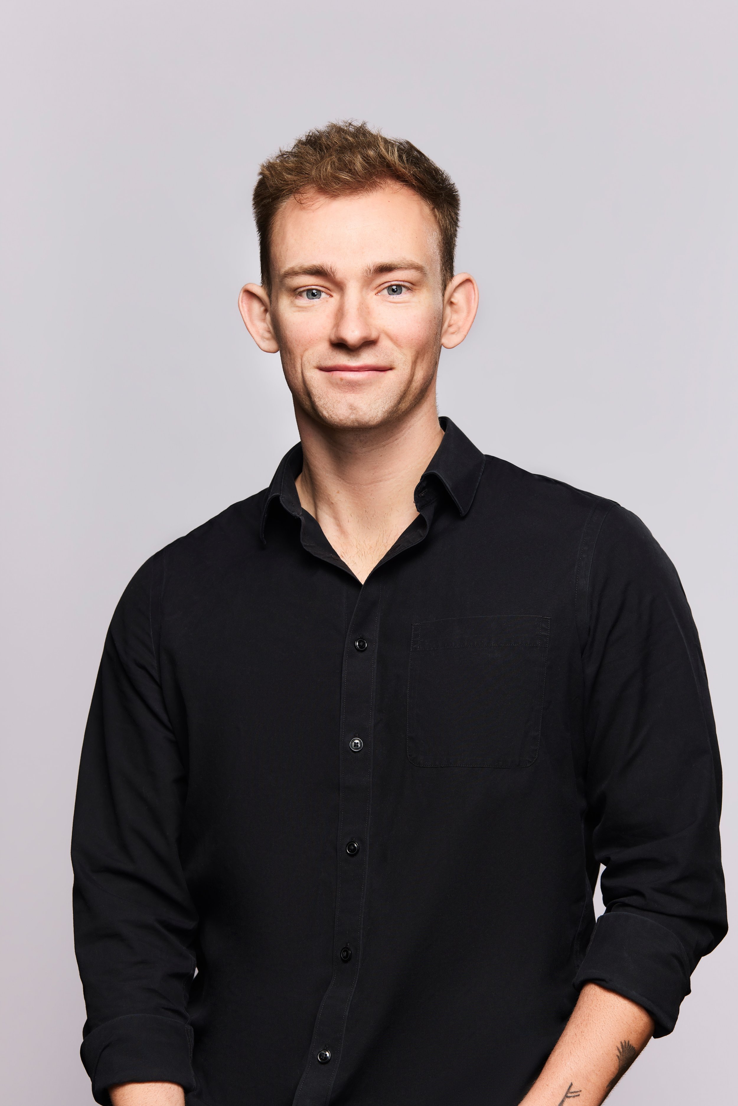 Portrait of a smiling man with short brown hair wearing a black button-up shirt against a light gray background