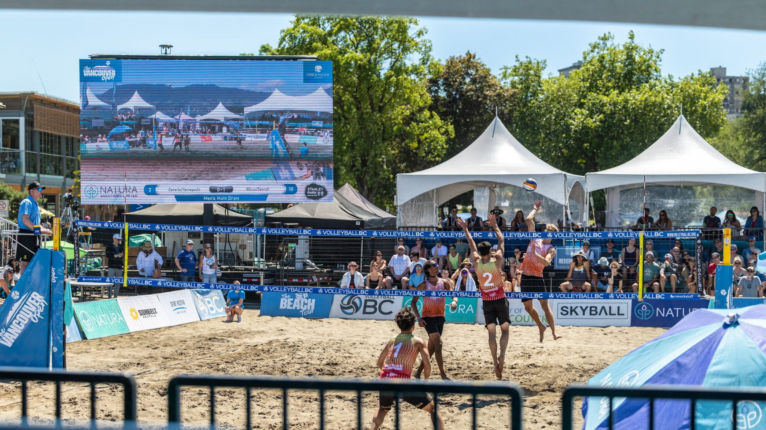 Beach volleyball players competing on a sand court at an outdoor tournament with spectators and a large scoreboard