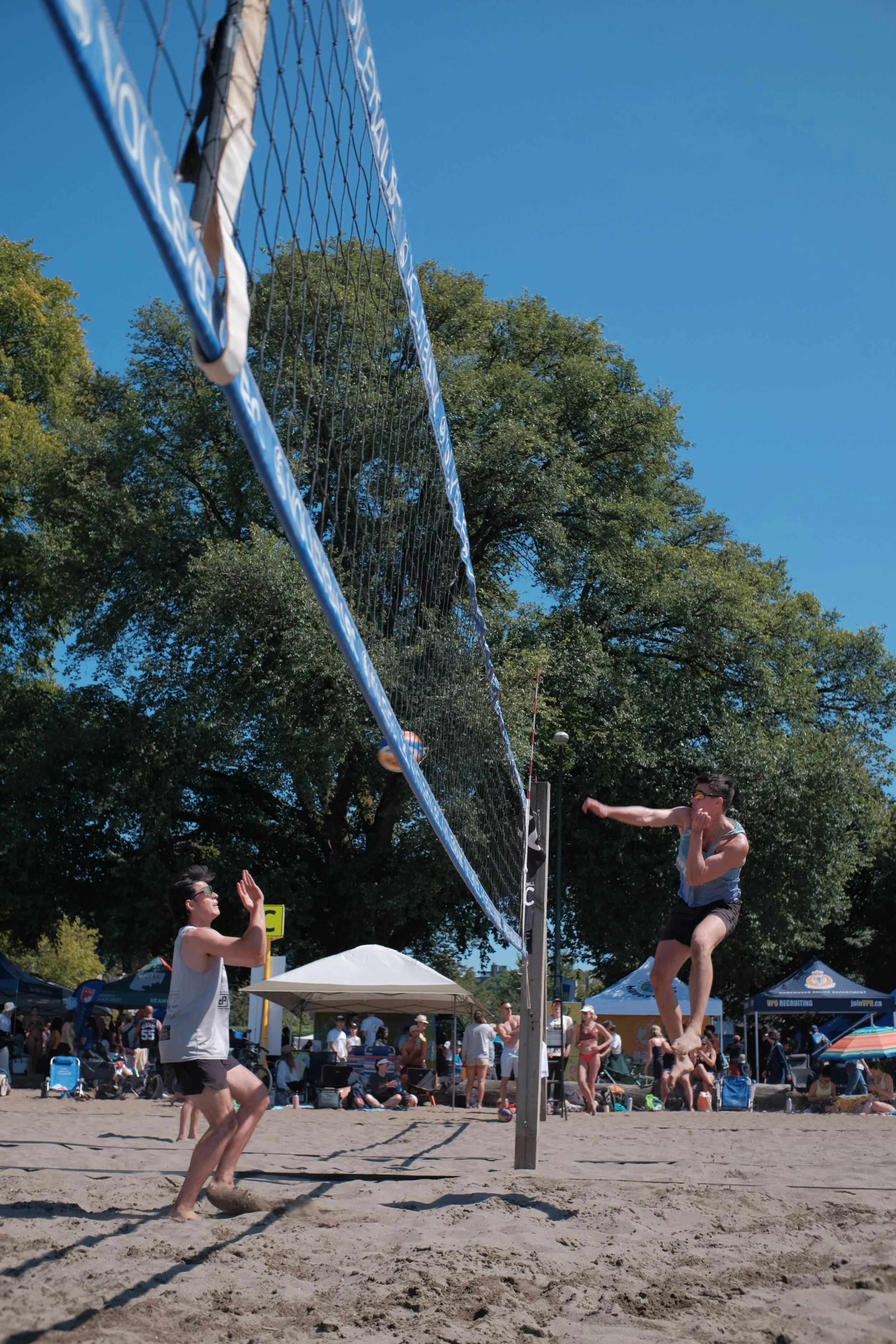 Beach volleyball players compete on a sandy court under a clear blue sky, with spectators and event tents in the background.