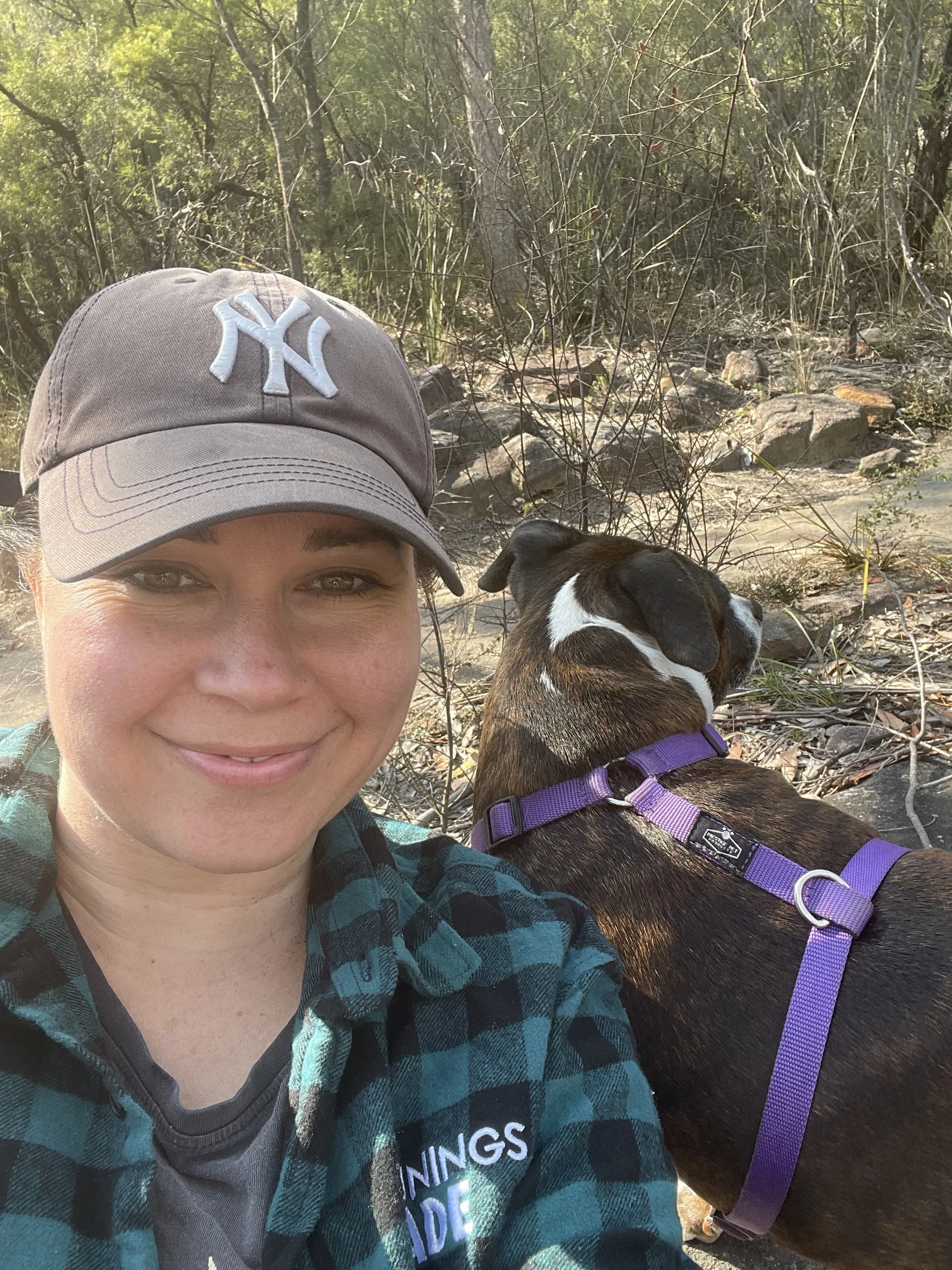 A woman wearing a grey New York Yankees baseball cap smiling outdoors with a small brown and white dog on a purple leash nearby in a wooded area.