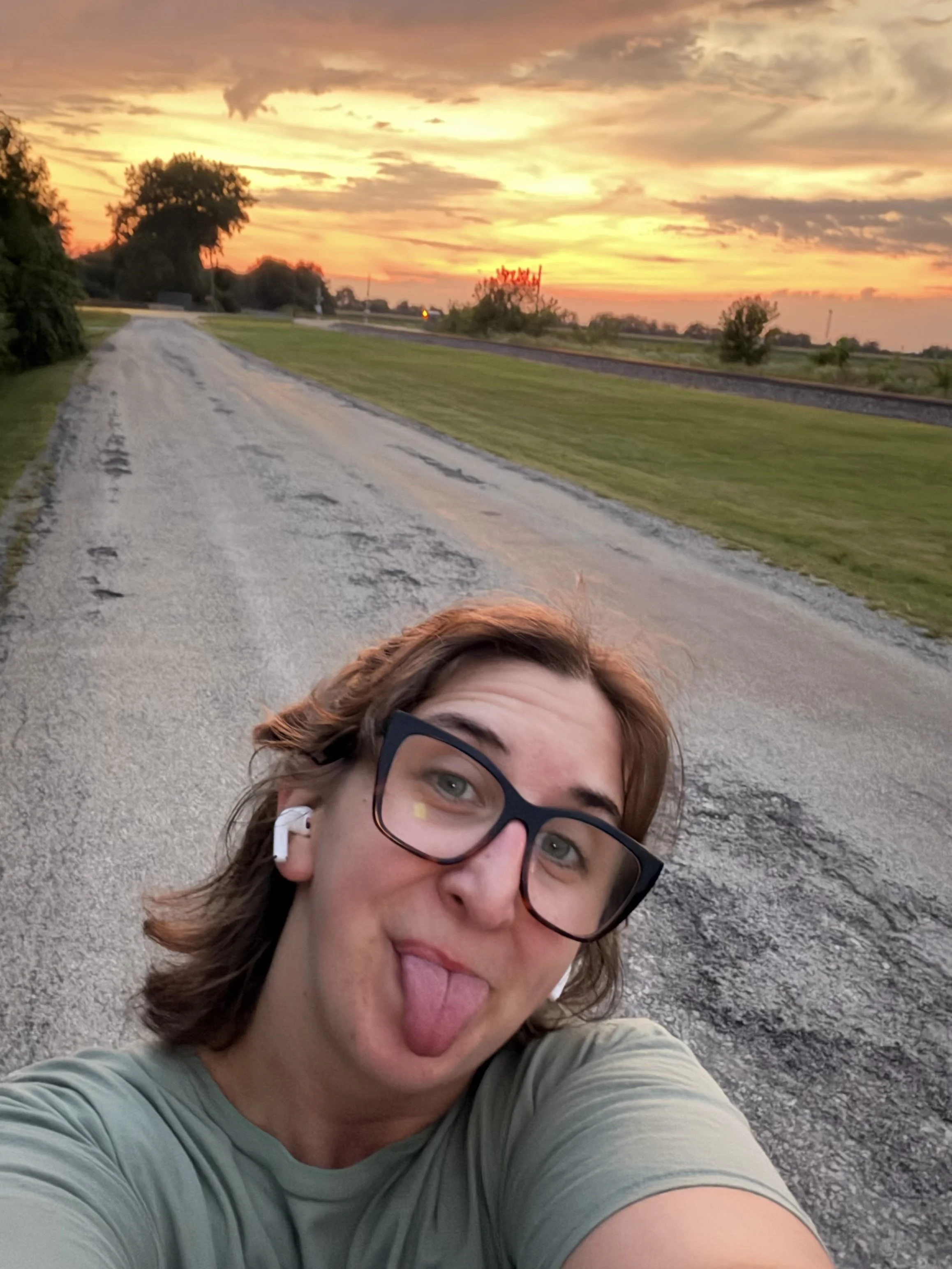 A woman with brown hair and glasses making a funny face while taking a selfie outdoors with a sunset sky and empty road in the background.