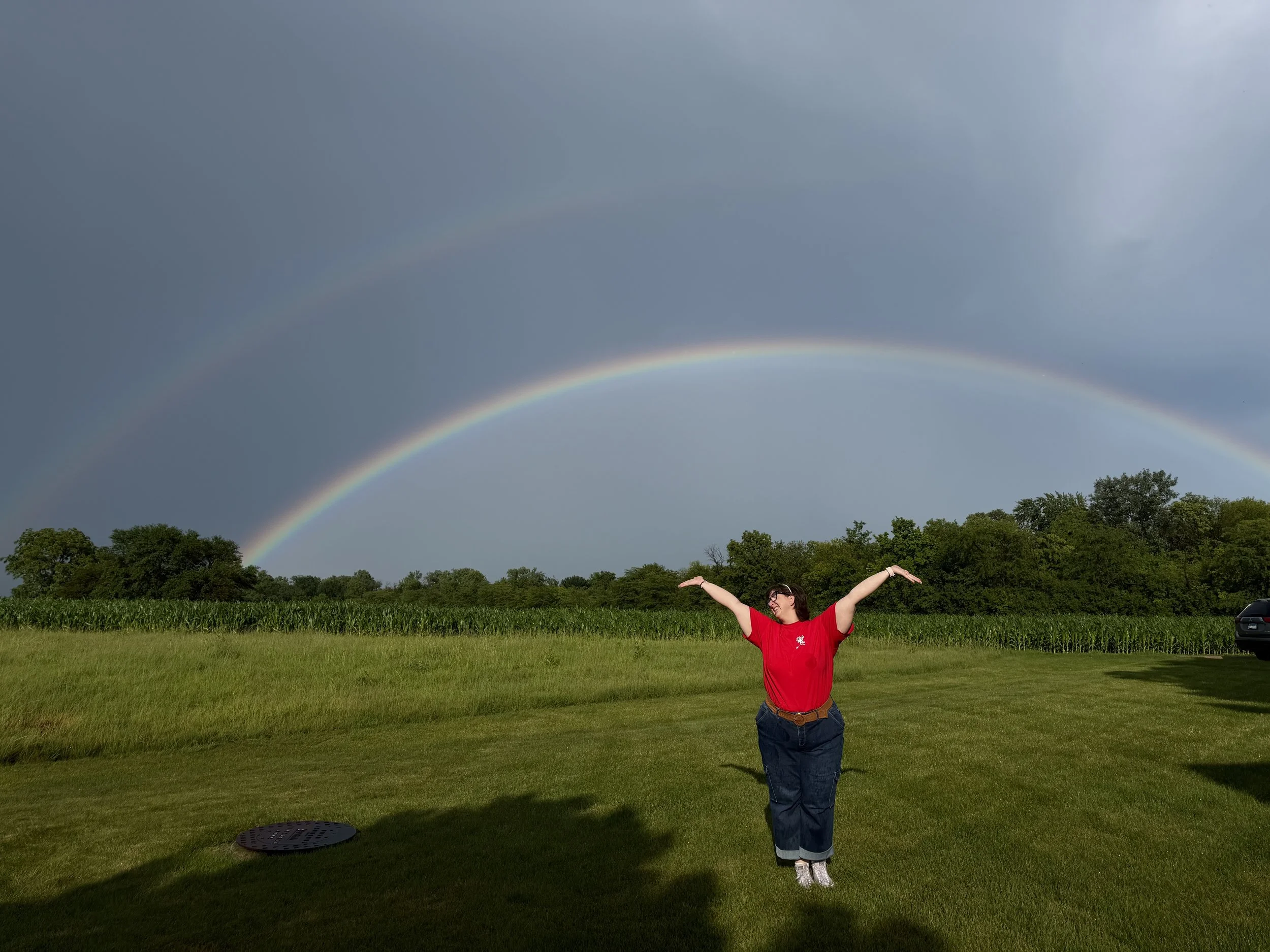 A woman standing in a grassy field with her arms raised, a rainbow in the sky behind her, over a backdrop of trees and a partly cloudy sky.