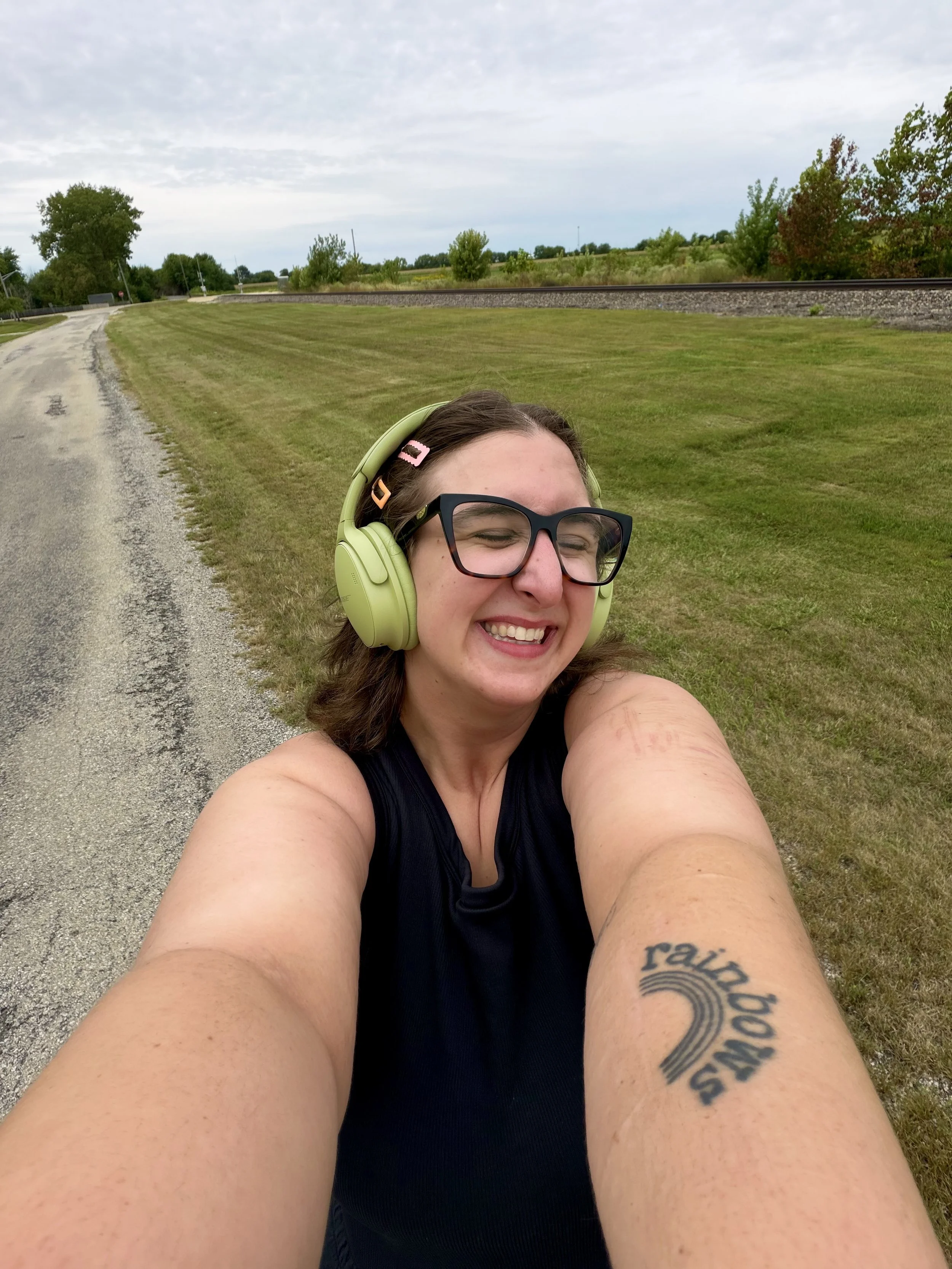 A smiling woman wearing glasses and green headphones taking a selfie on a grassy area next to a railroad track.