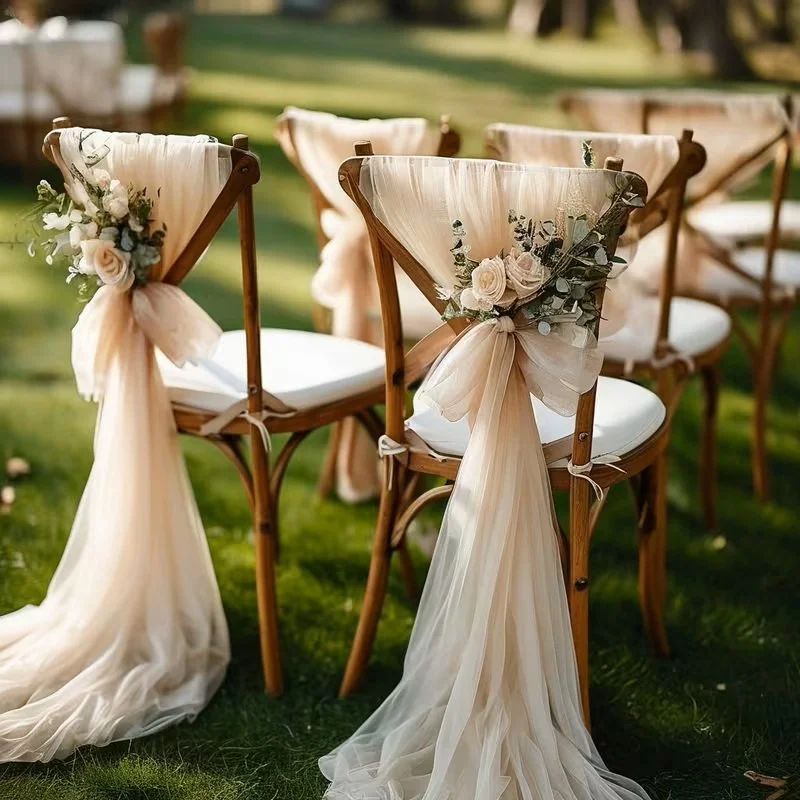 Wedding chairs decorated with cream-colored tulle and floral arrangements, placed outdoors on a grassy area.