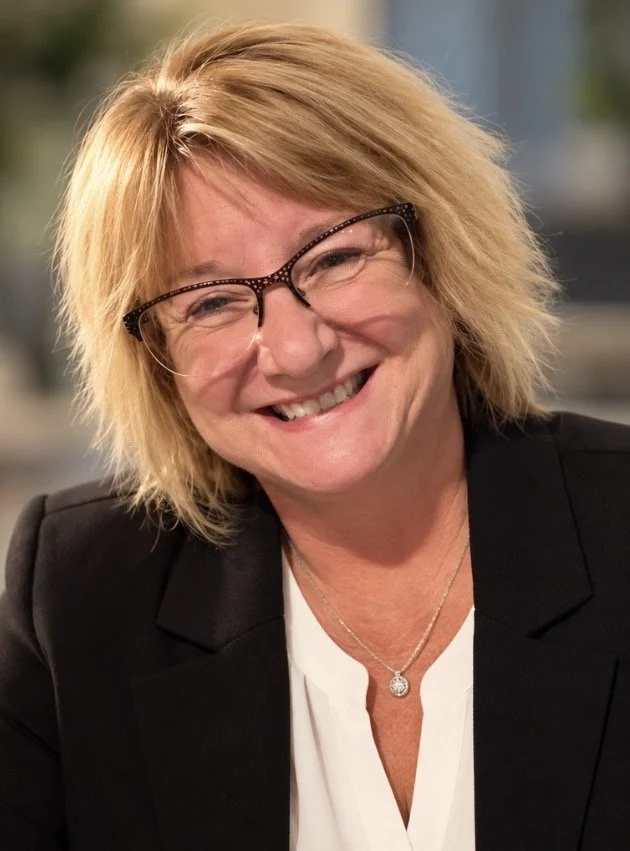 A woman with blonde, tousled hair and glasses, smiling at the camera. She is wearing a black blazer over a white top and a silver necklace with a round pendant.