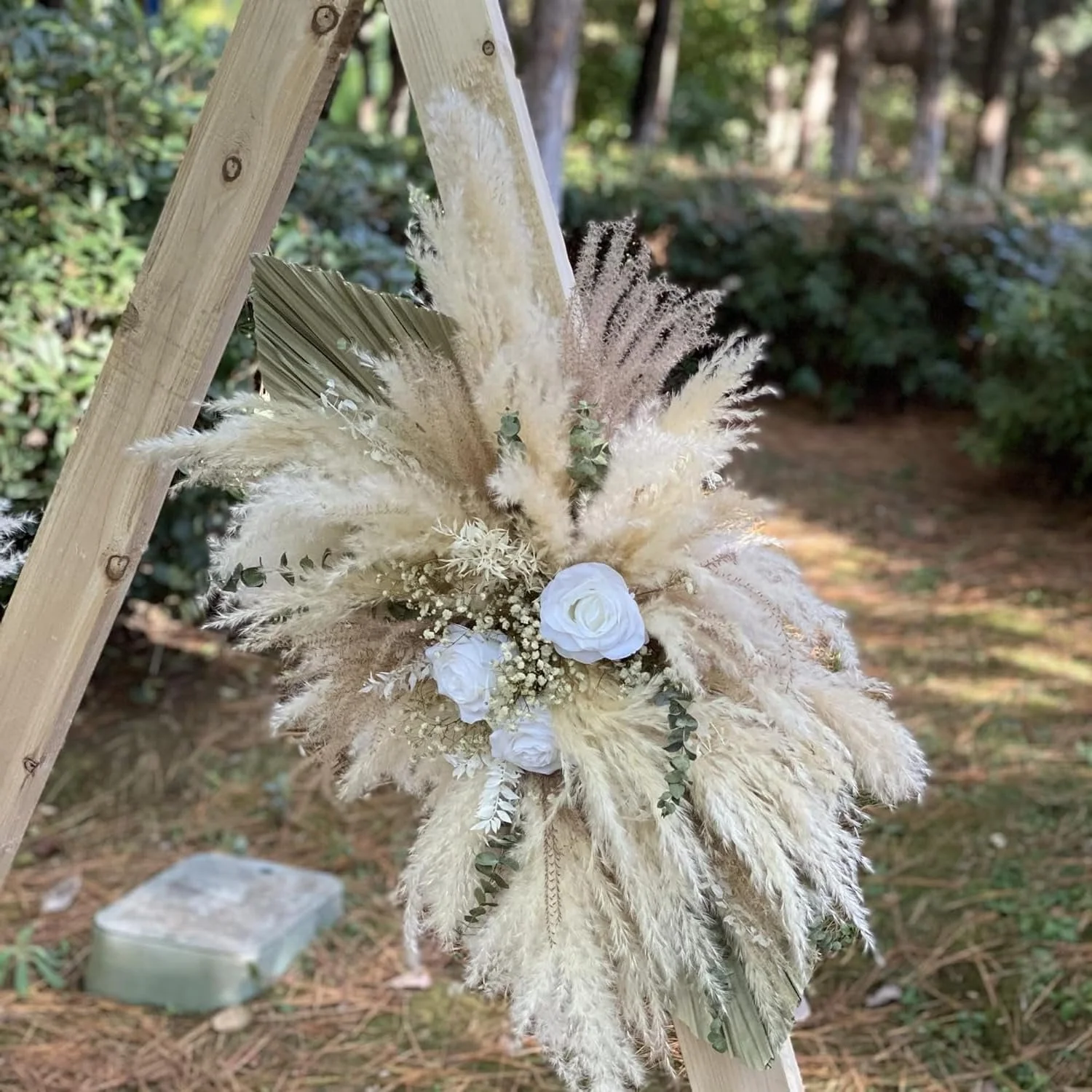 A floral arrangement with white flowers and beige pampas grass, attached to a wooden structure outdoors in a wooded area.