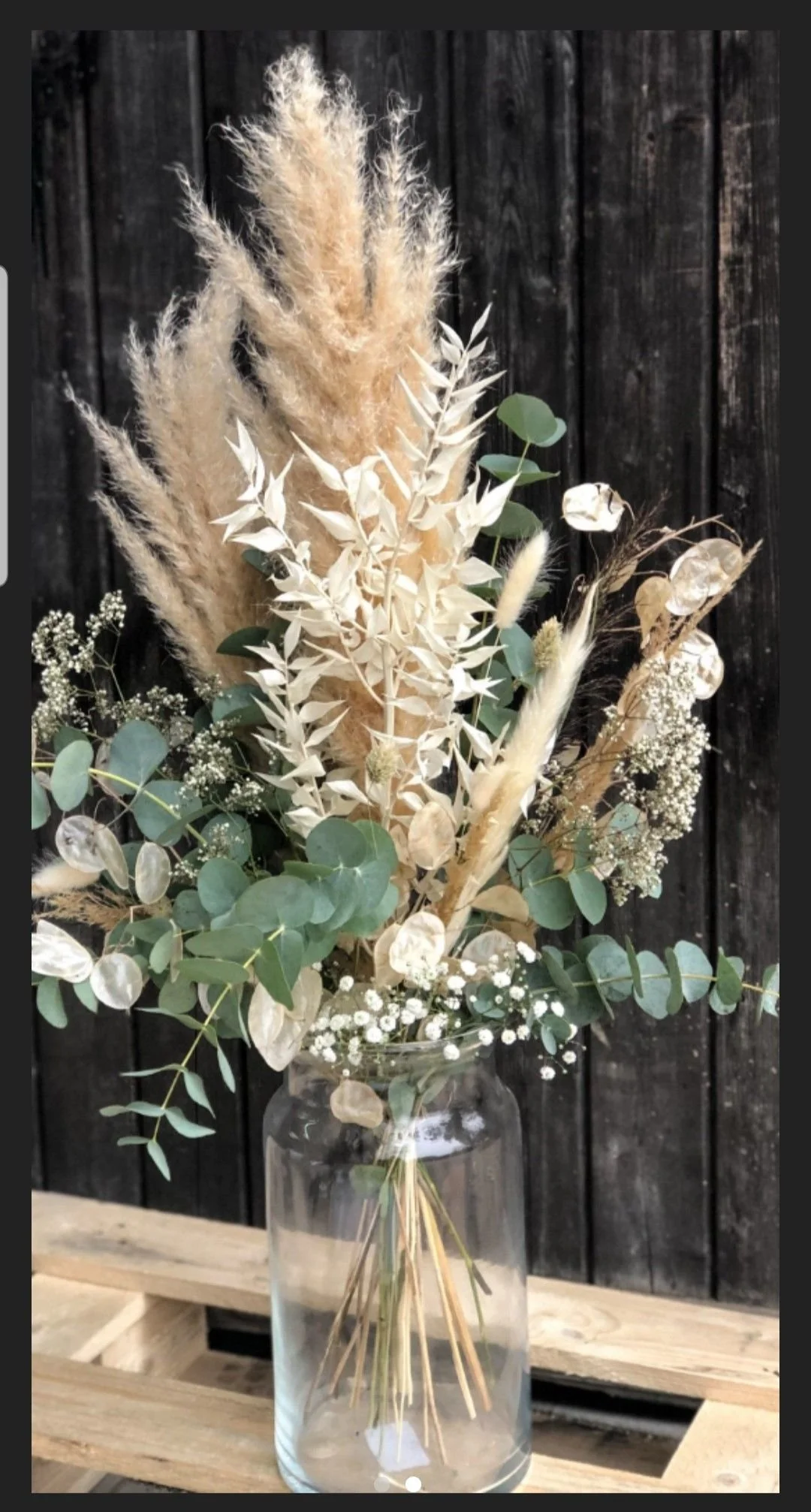 A bouquet of dried flowers and greenery in a clear glass vase on a wooden surface, with a black wooden wall background.